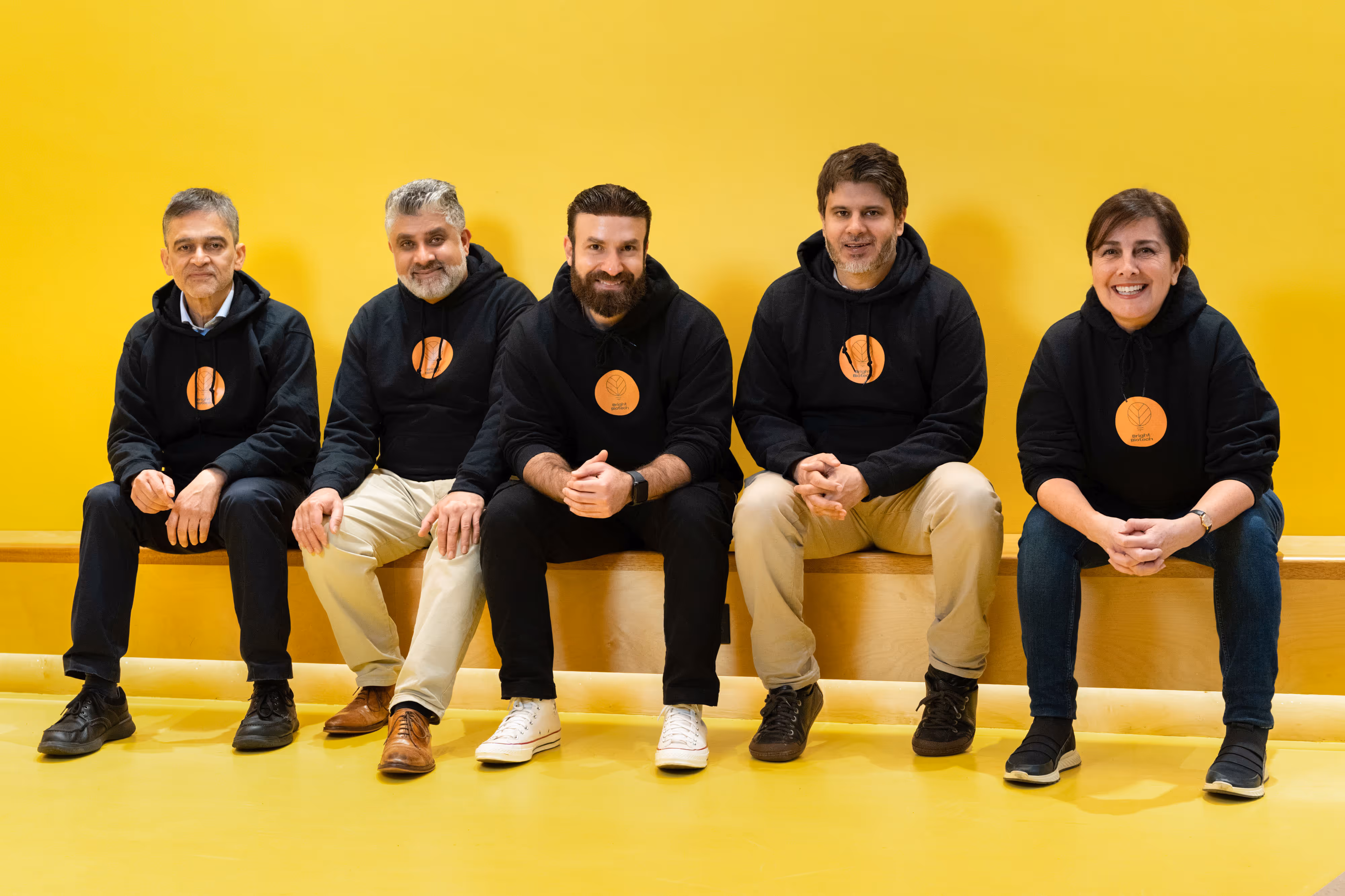 Five people from a biotech company pose in black hoodies sit and smile against a bright yellow wall and floor.