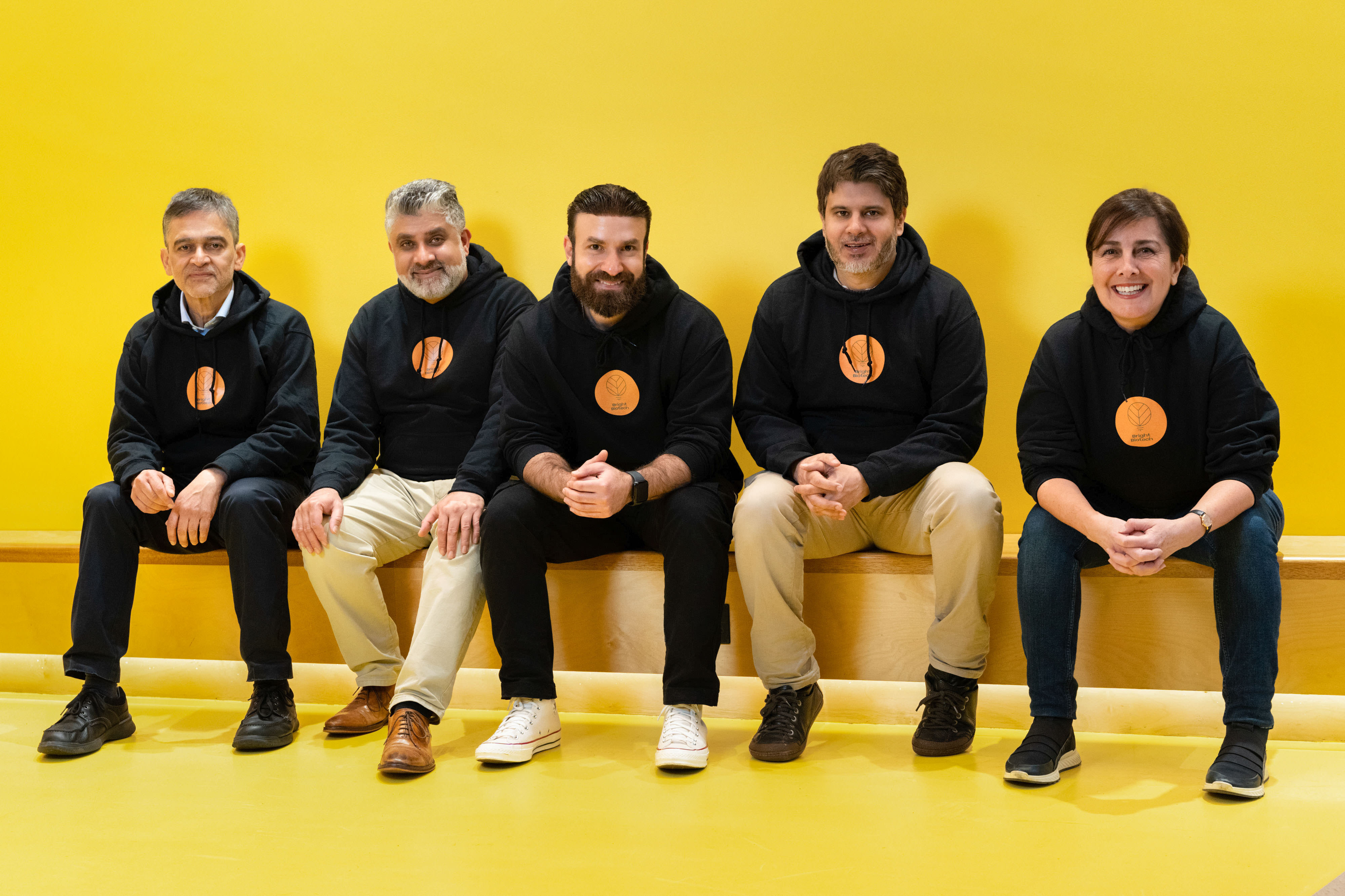 Five people from a biotech company pose in black hoodies sit and smile against a bright yellow wall and floor.
