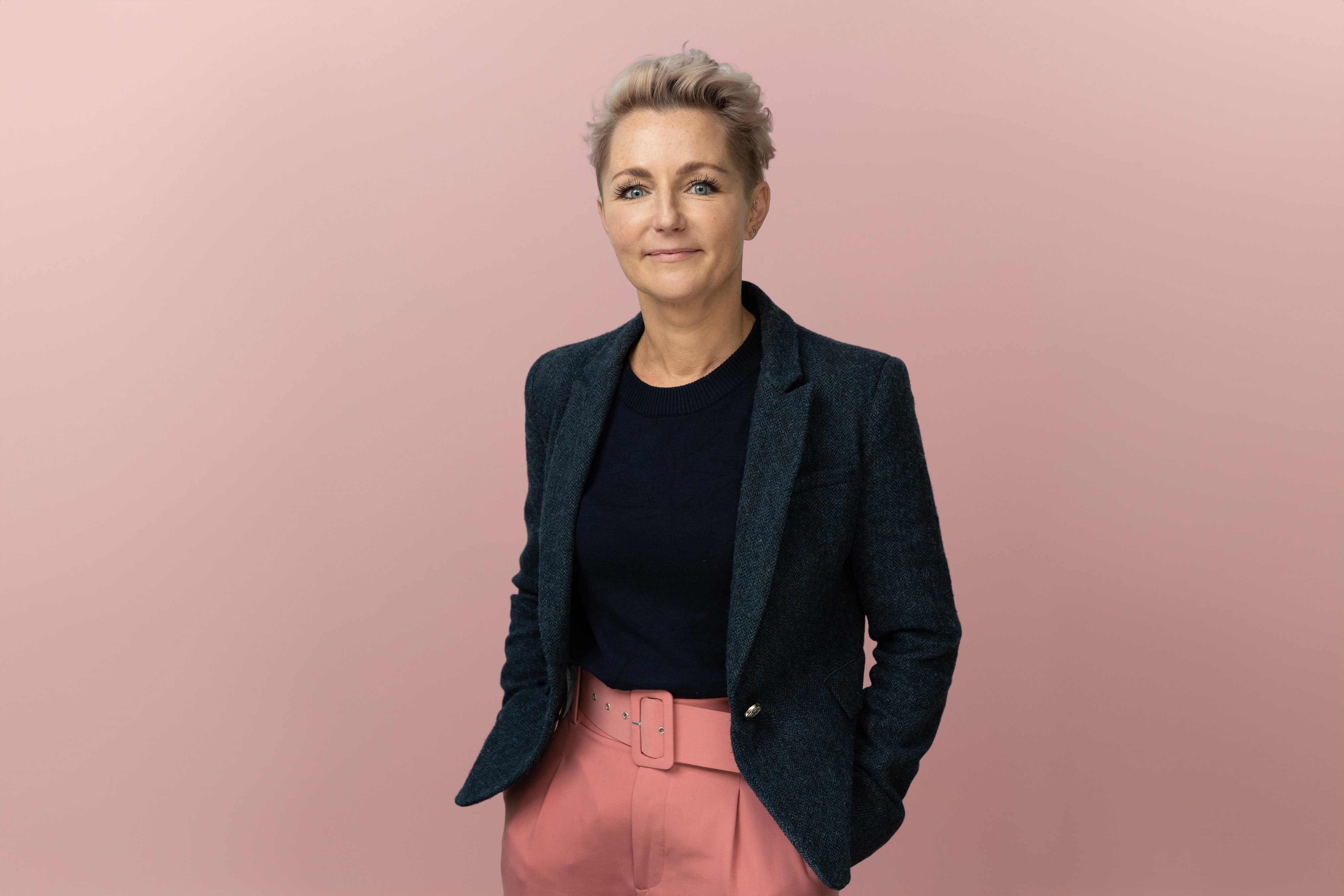 Confident person with short hair in a blazer and pink trousers stands against a plain pink background, hands in pockets.