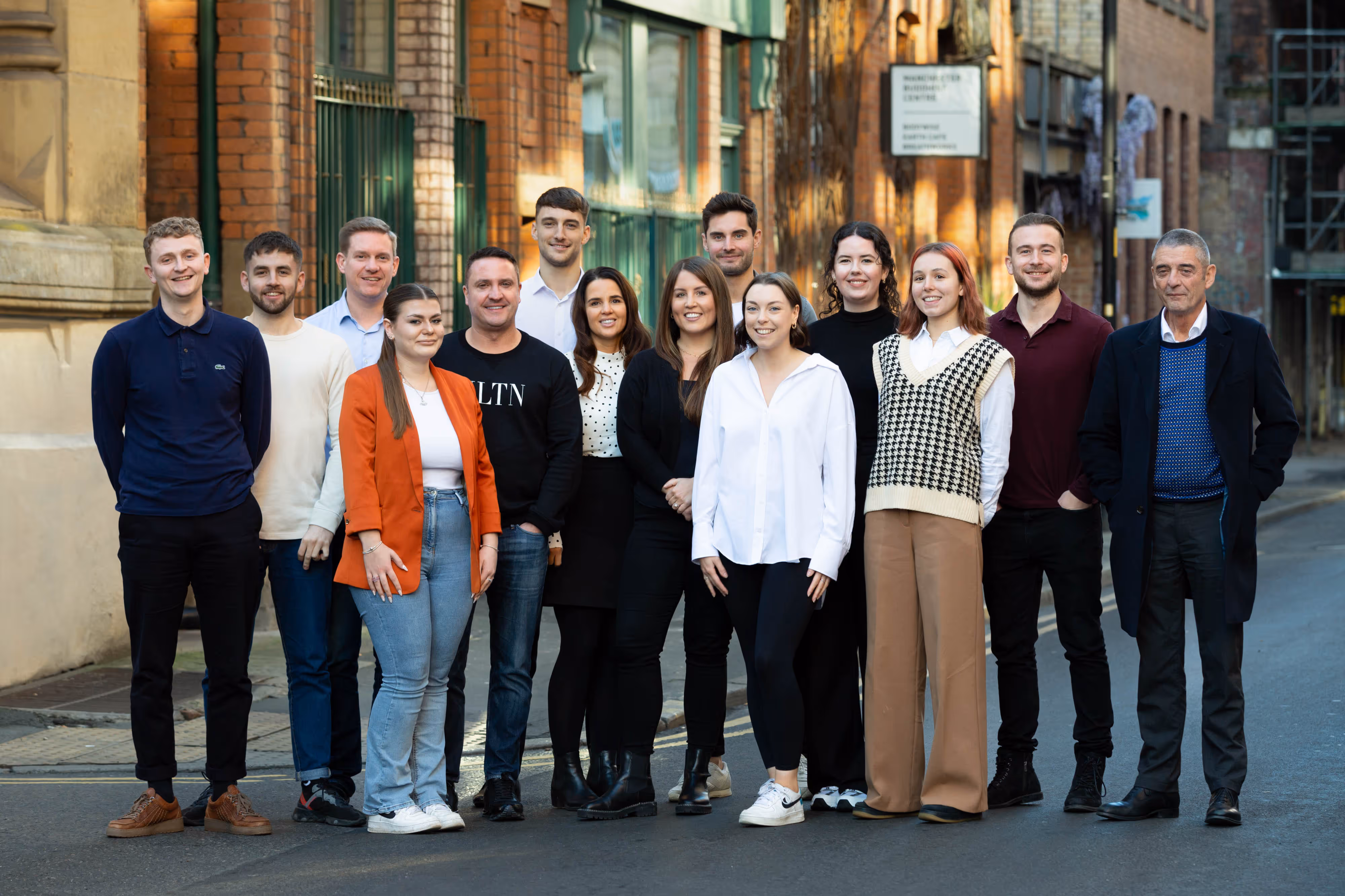 A group of fourteen people standing and smiling together outdoors on a city street in the Northern Quarter, Manchester.
