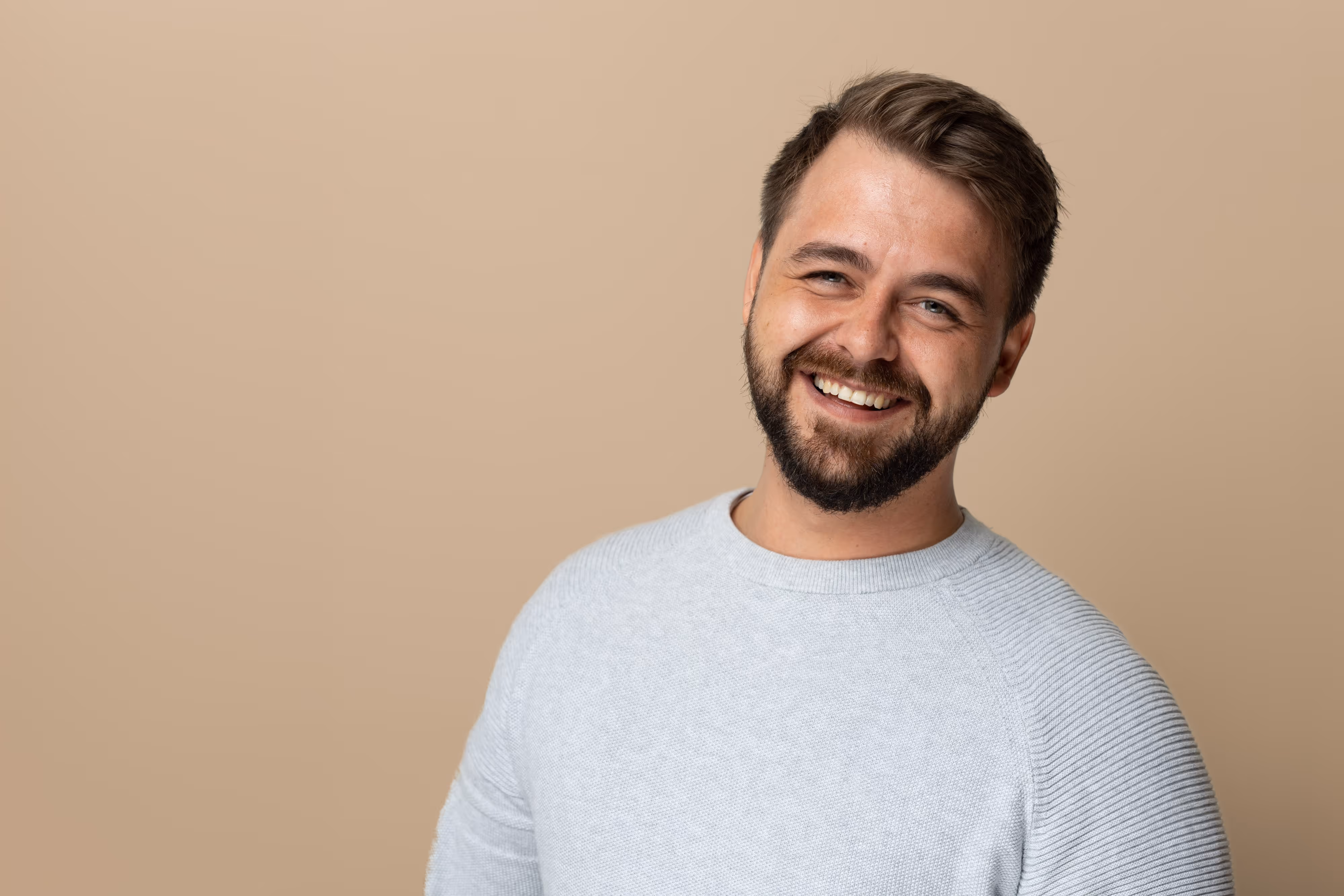 Smiling staff member with a beard wearing a light grey jumper standing against a plain beige background.