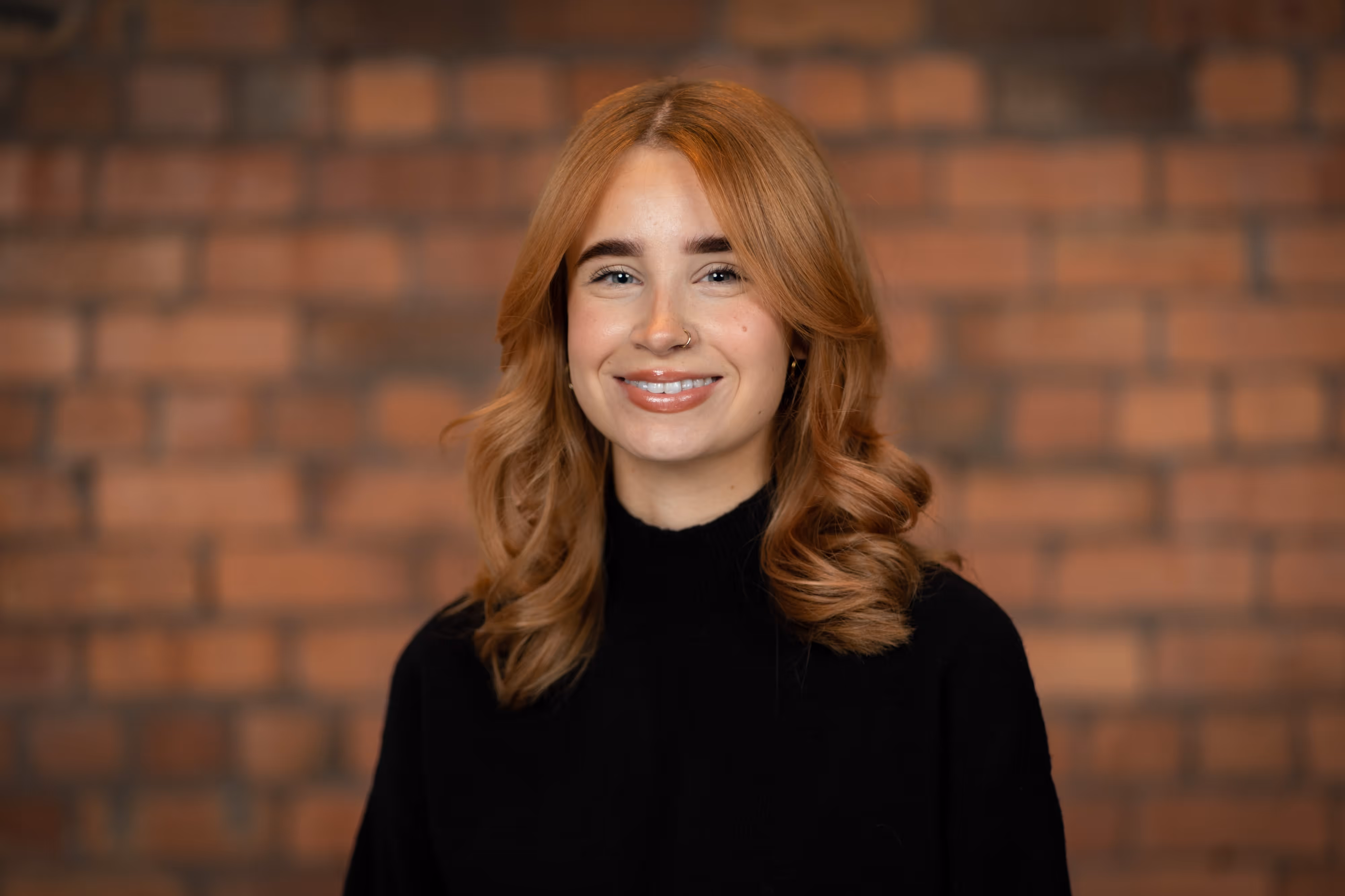 A smiling woman with wavy auburn hair wearing a black top stands in front of a brick wall posing for a staff headshot.