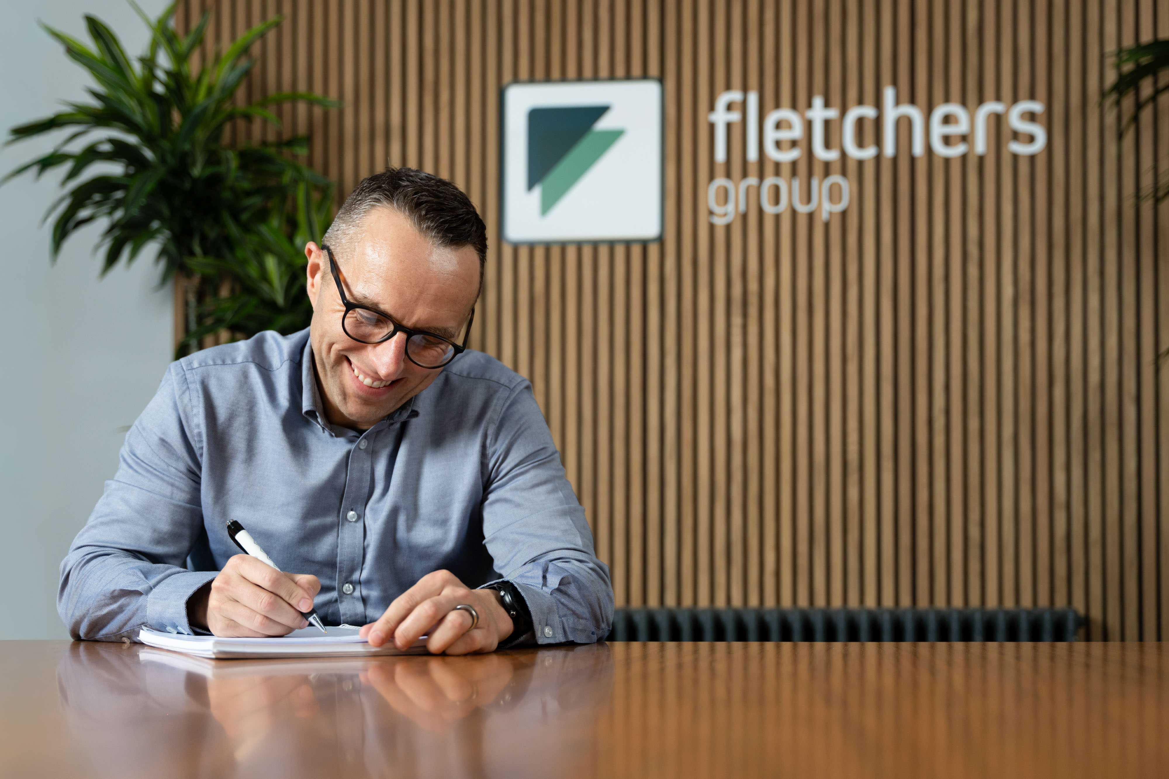 Smiling man in glasses writes on paper at a table with "fletchers group" logo on wood panelled wall behind him.