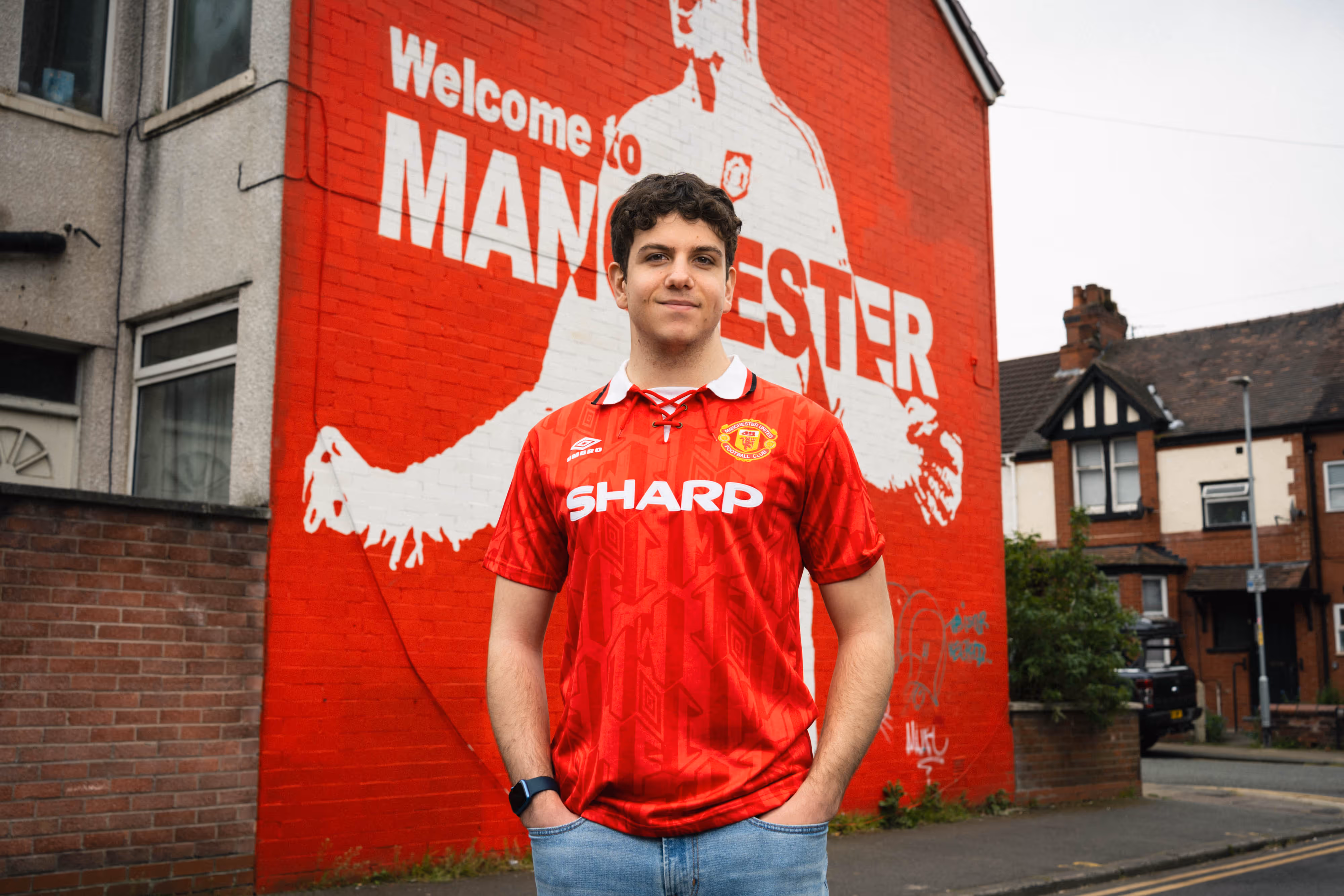 Young man in a red Manchester United shirt stands in front of a red mural that says "Welcome to Manchester".