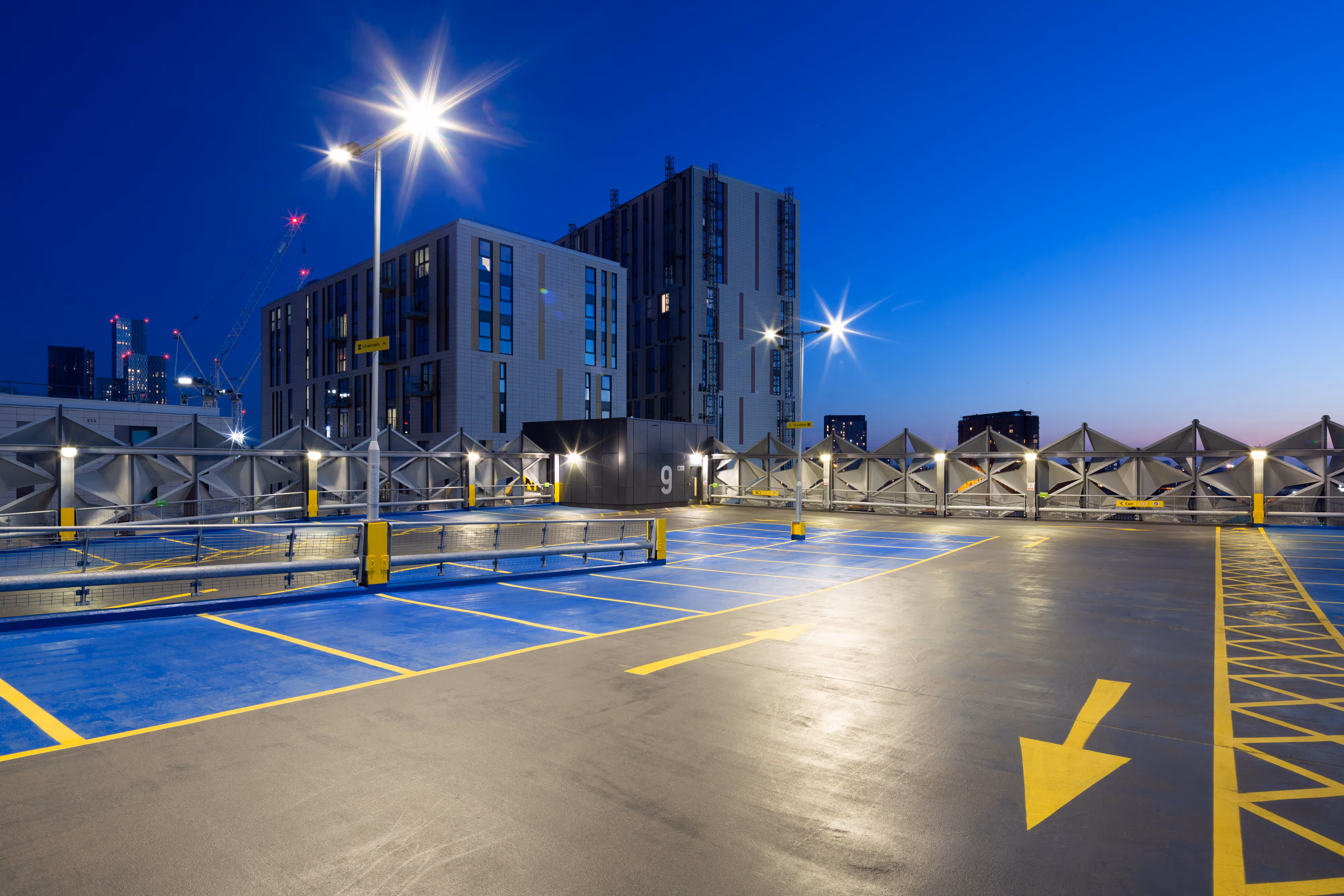 Empty rooftop car park at dusk with blue spaces, yellow arrows, and modern buildings in the background.