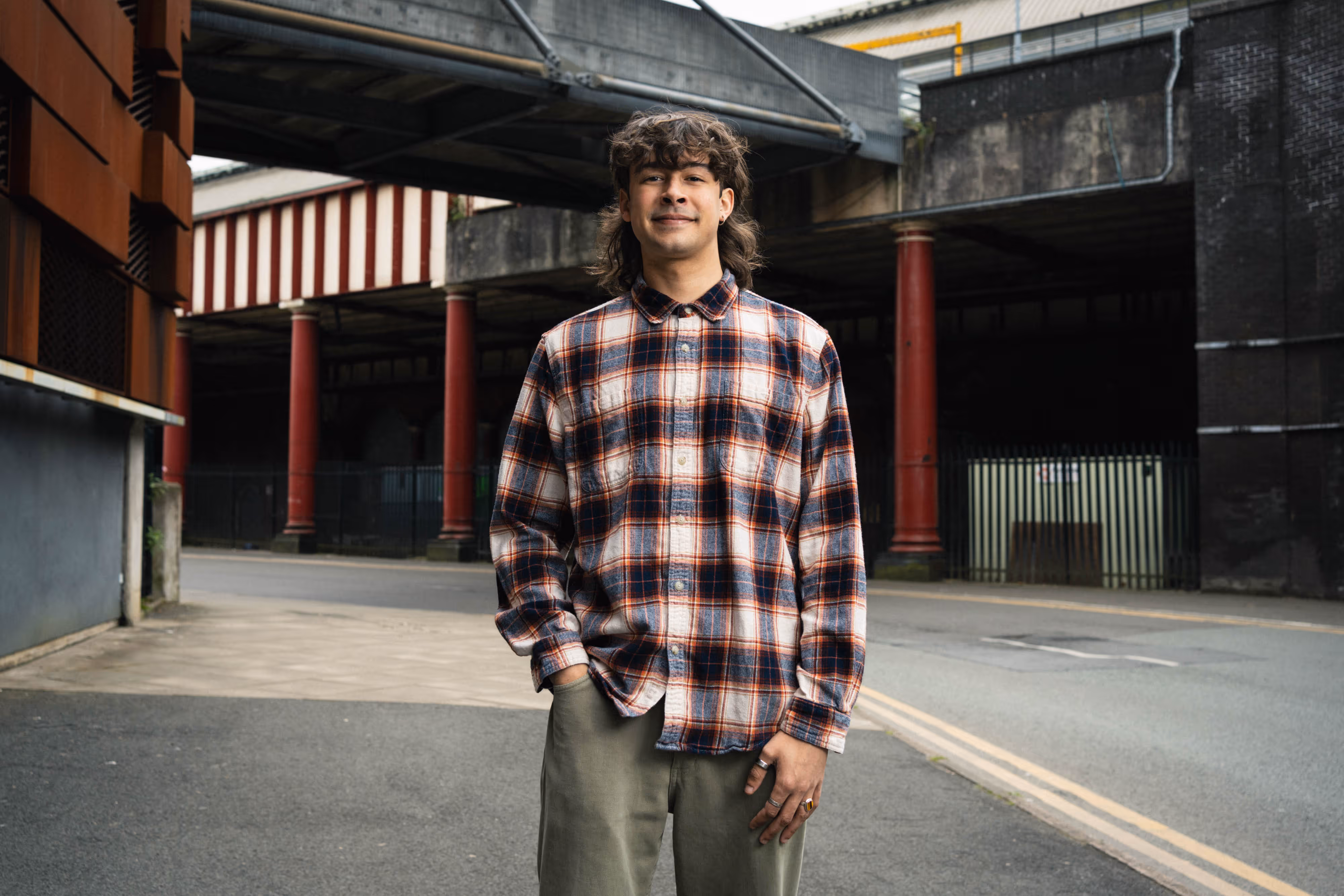 A Manchester University student with curly hair in checked shirt standing on an empty urban street under a flyover, smiling slightly.