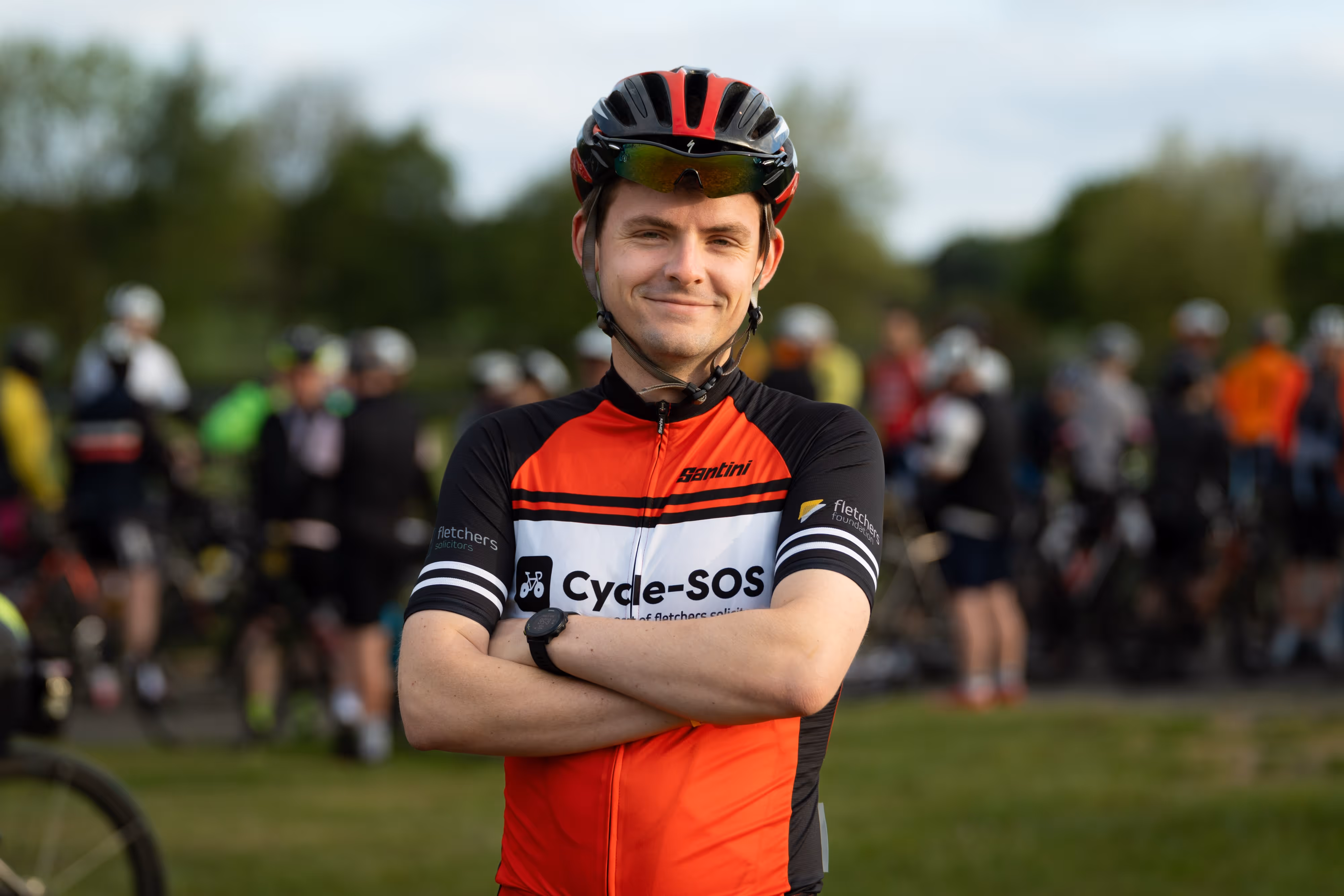 Smiling cyclist in a helmet and a branded red jersey stands with arms folded, with a group of cyclists in the background.