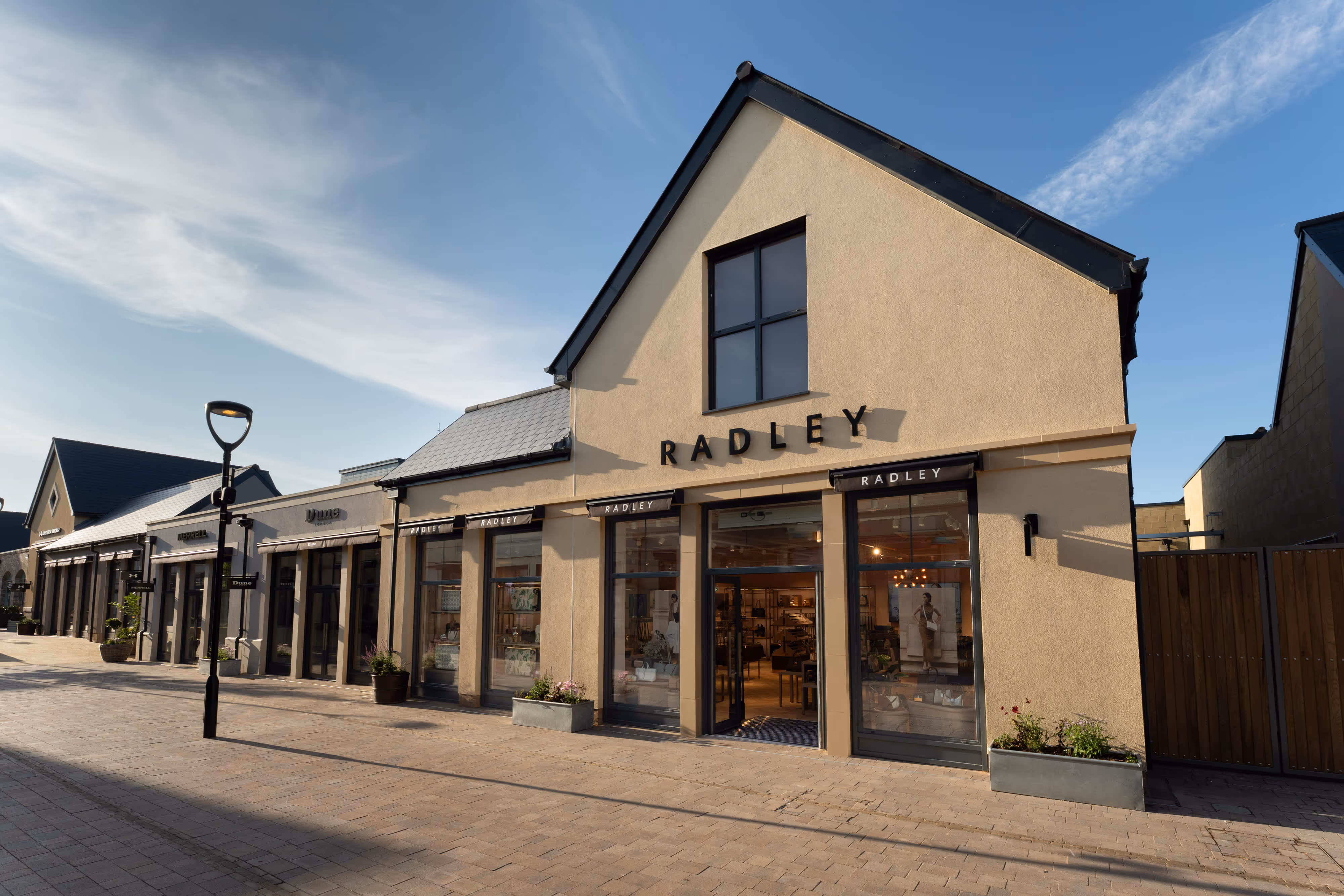A modern beige shopfront with large windows and "RADLEY" signage on a quiet, sunlit designer outlet.