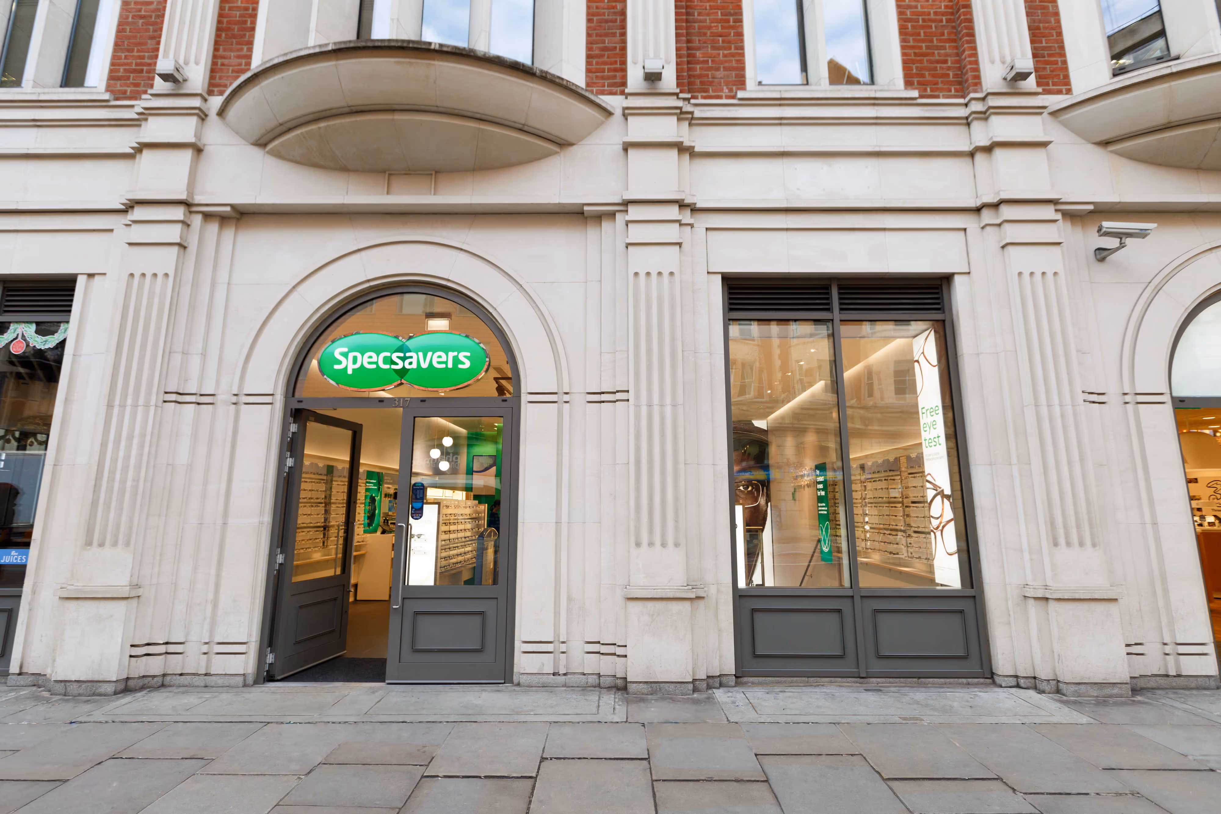 Specsavers shopfront with arched entrance, large windows, and illuminated green sign on a stone building.