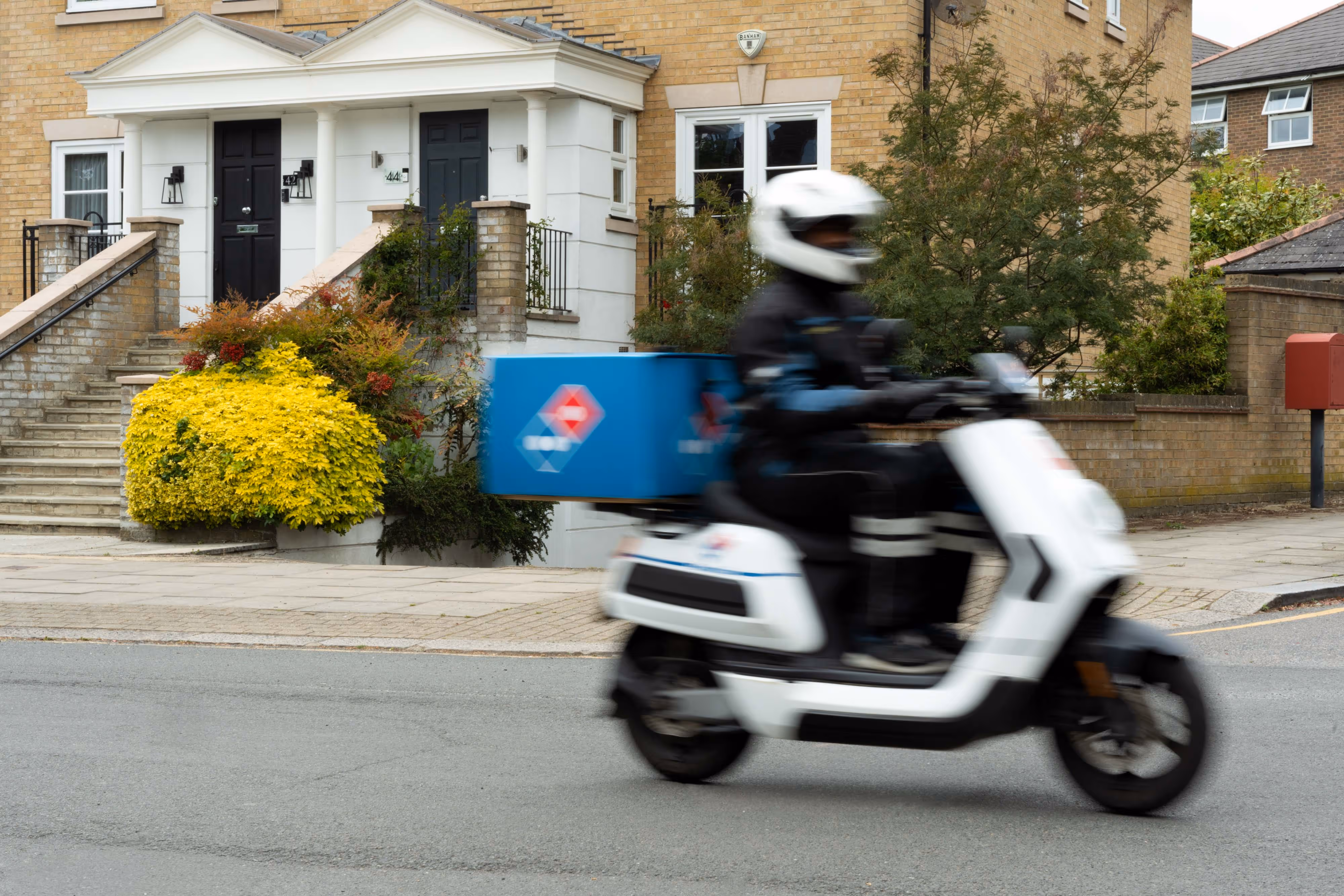 A pizza delivery person rides a scooter past houses on a residential road, captured with a slow shutter speed to show motion.