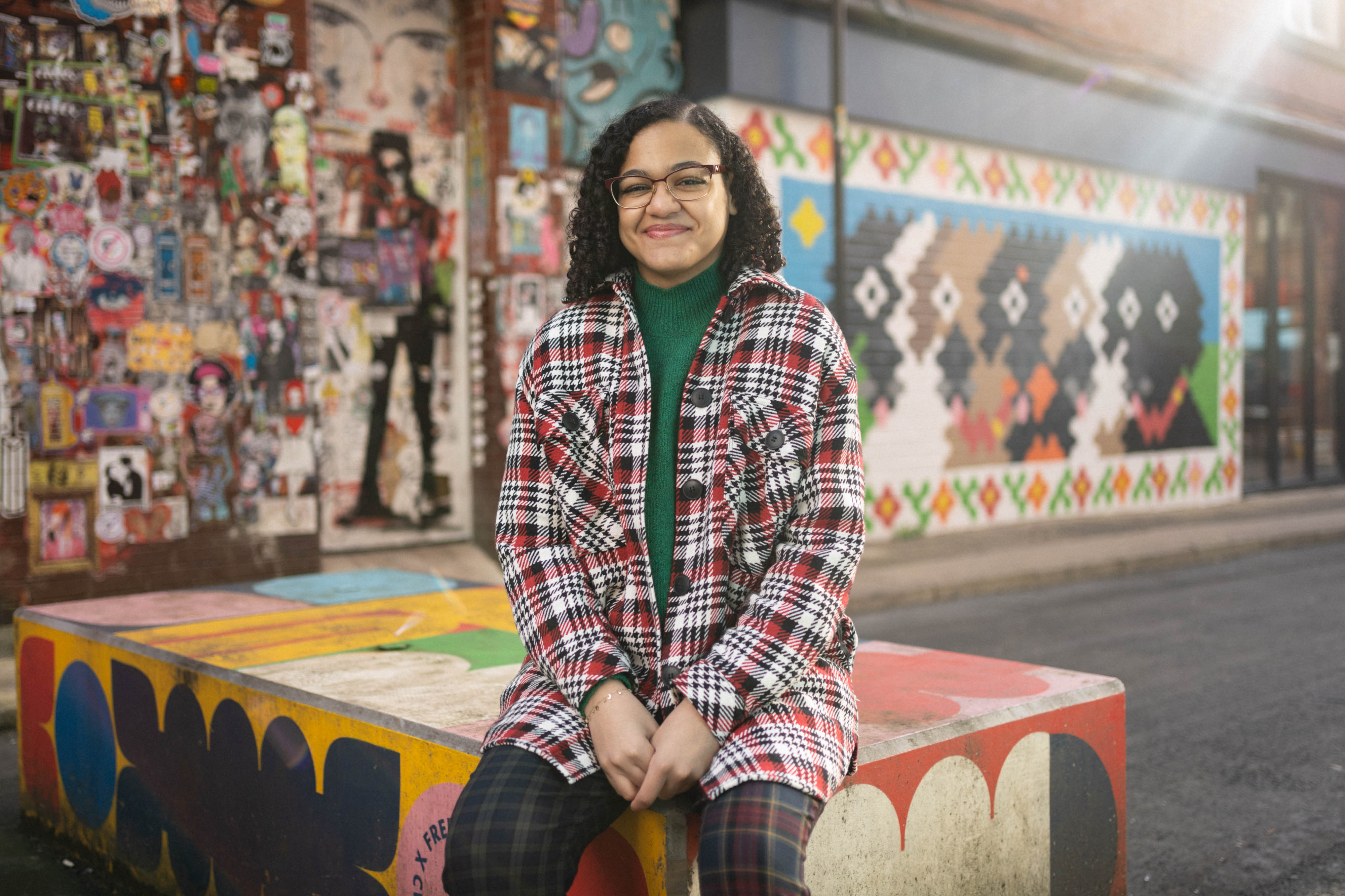 A smiling student in glasses and a checked coat sits on colourful street art with vibrant murals in the background.