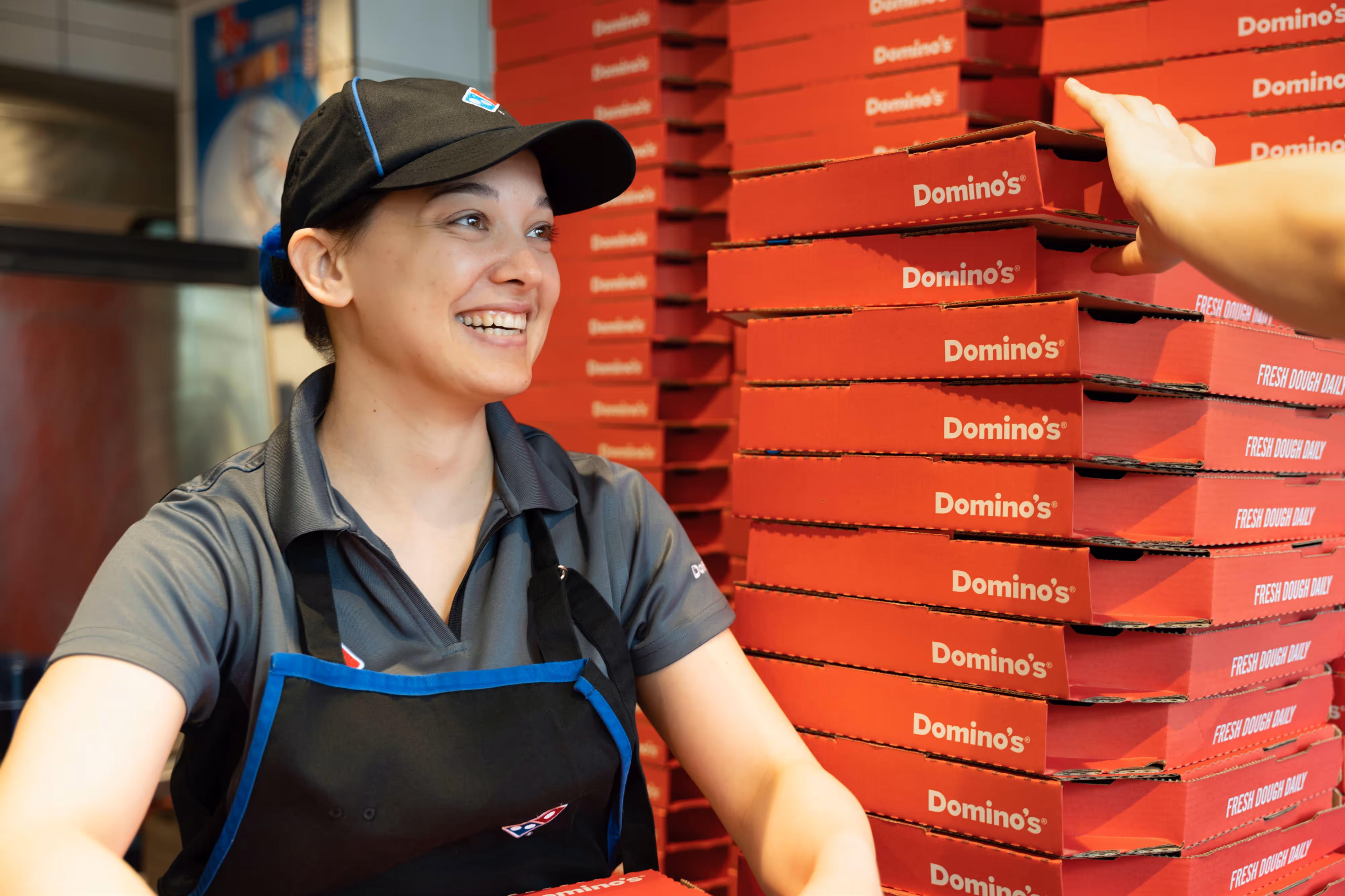 Smiling pizza restaurant worker in uniform next to a tall stack of pizza boxes, handing one to a customer.