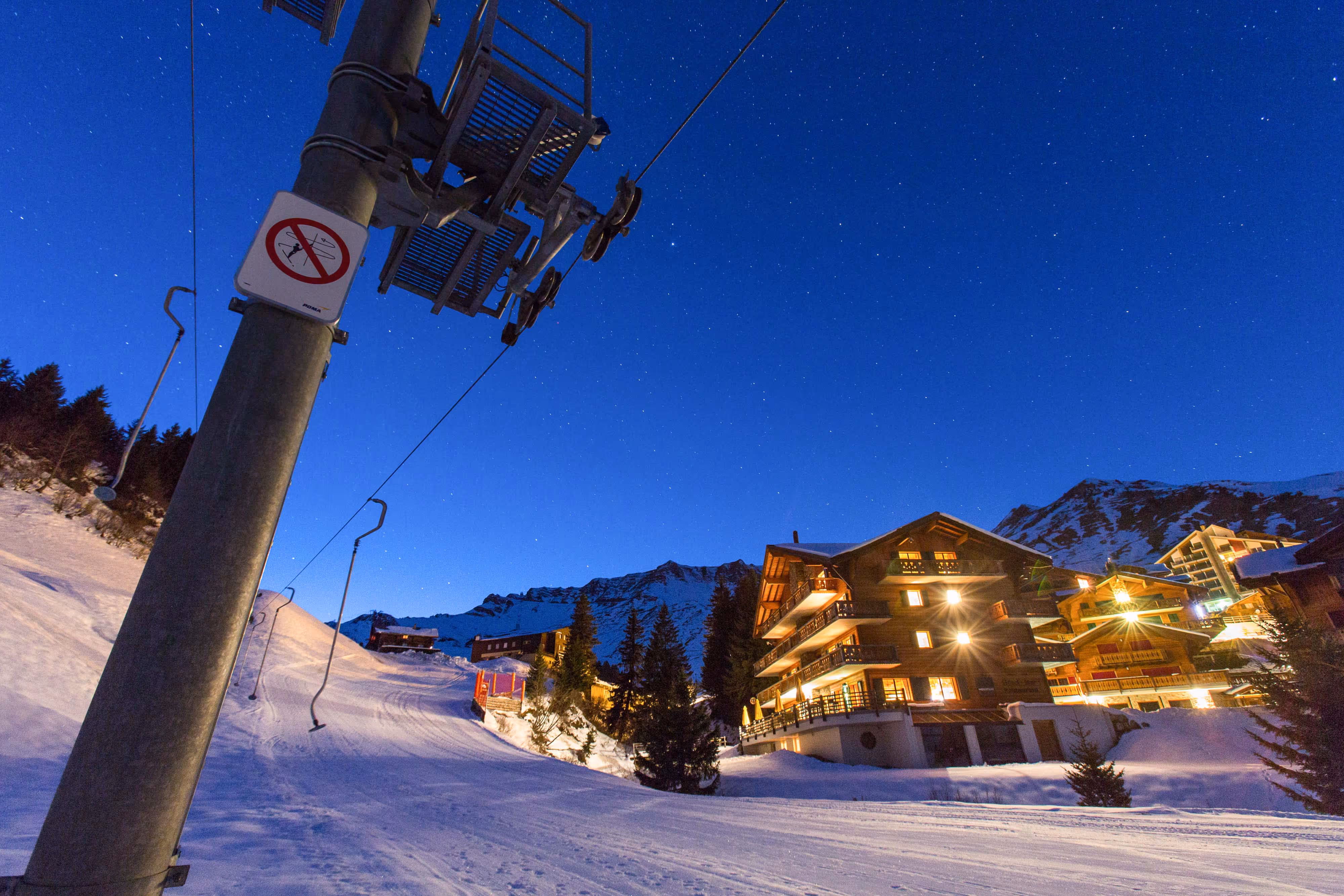 Ski resort buildings lit at night, snowy pistes, ski lift, and clear starry sky in a mountain setting.