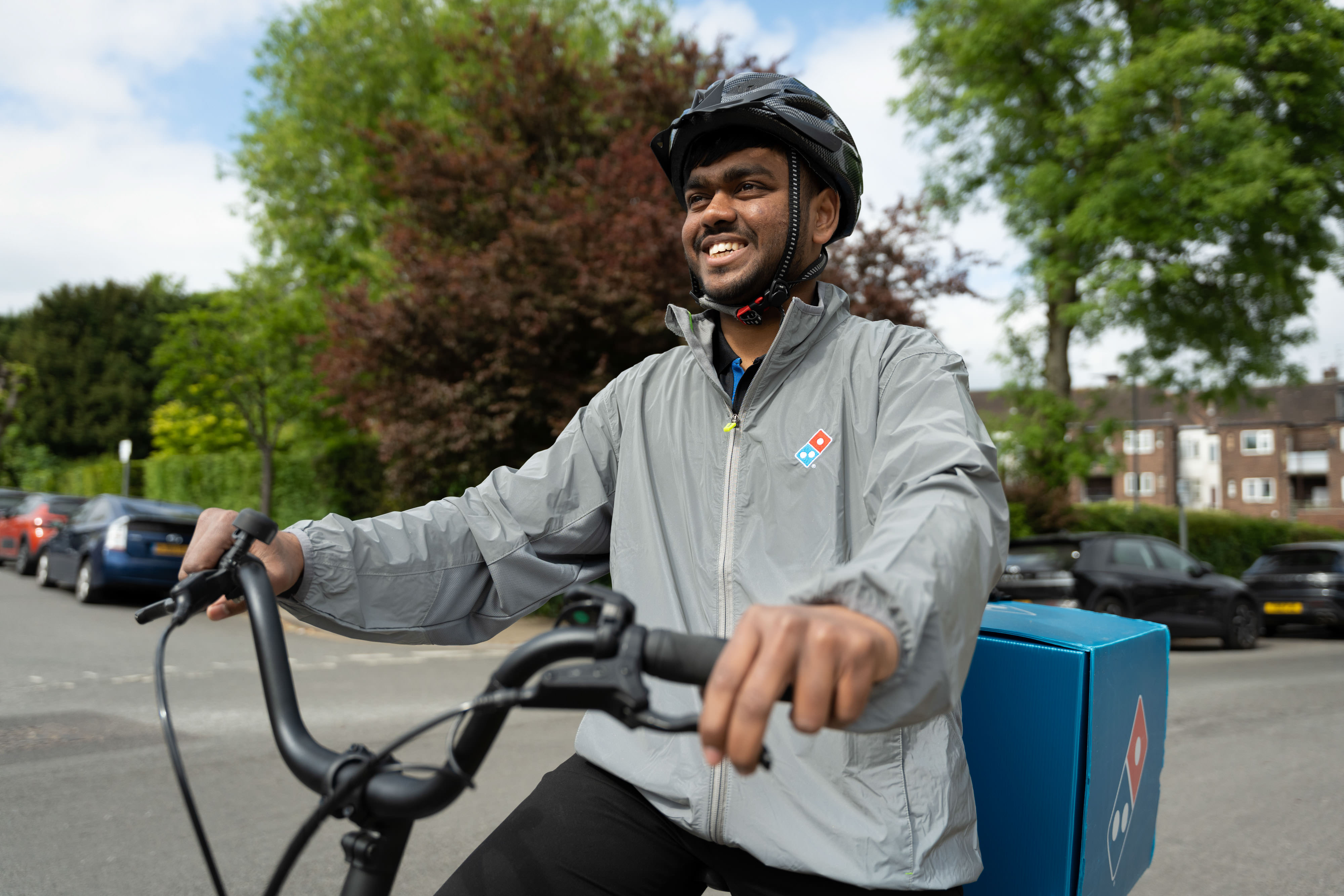 A smiling pizza delivery person rides a bicycle on a street, wearing a helmet and high vis grey jacket.