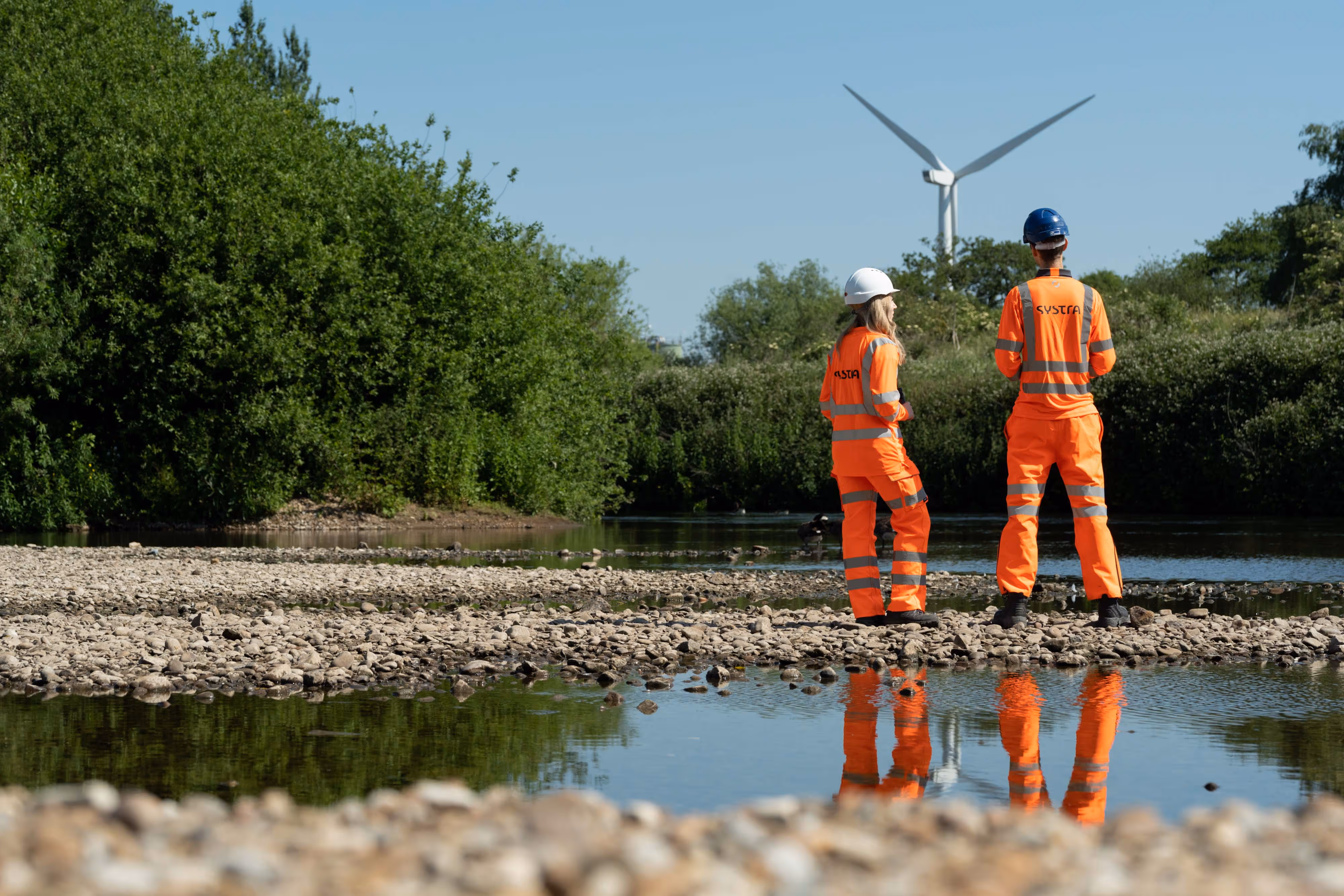 A couple of environmental workers in orange uniforms surveying a nearby wind turbine.