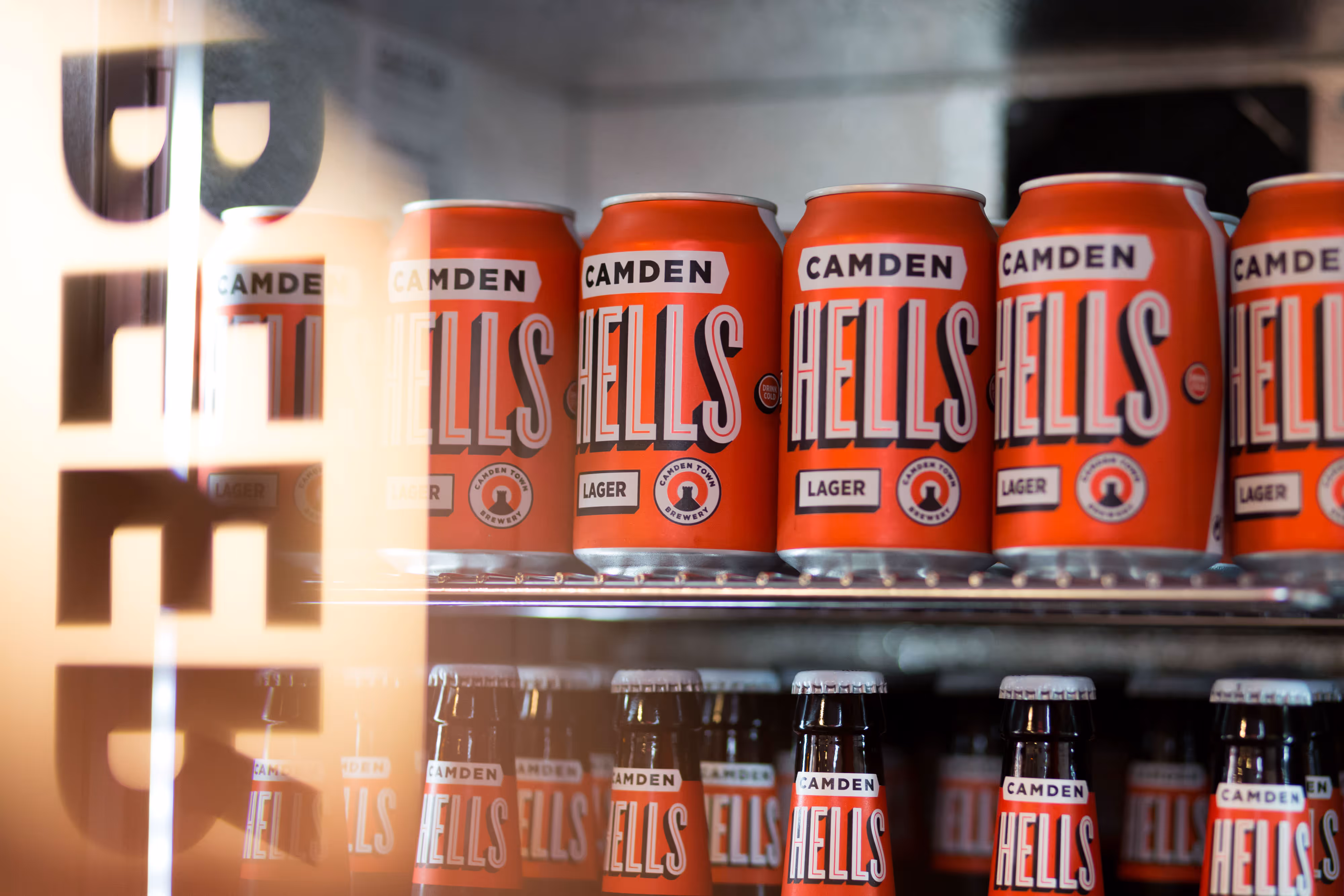 Cans and bottles of Camden Hells Lager on a shelf in a fridge, with "BEER" reflected on the glass door.