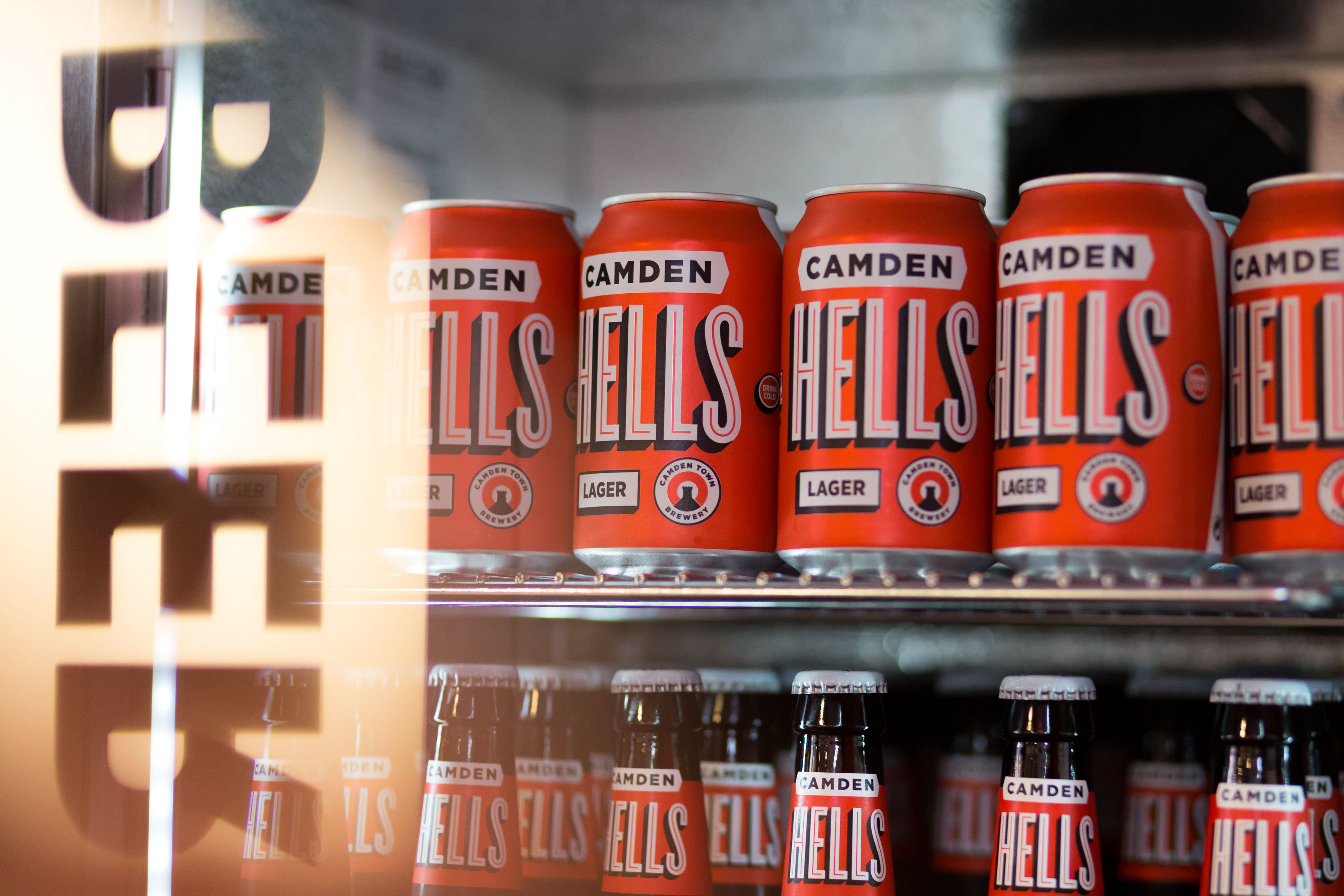 Cans and bottles of Camden Hells Lager on a shelf in a fridge, with "BEER" reflected on the glass door.