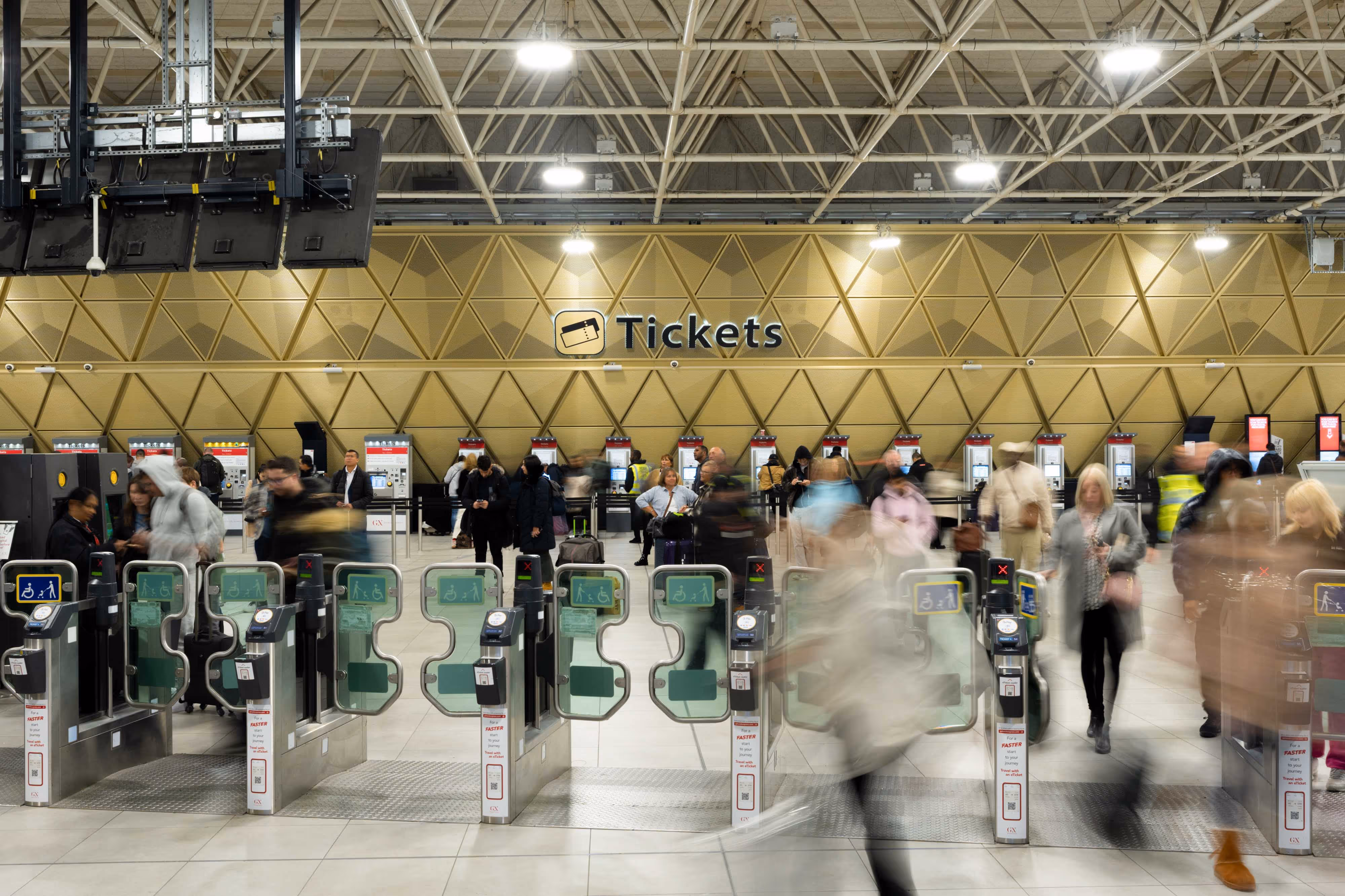 People passing through ticket barriers at a busy railway station with ticket machines and a "Tickets" sign in the background.