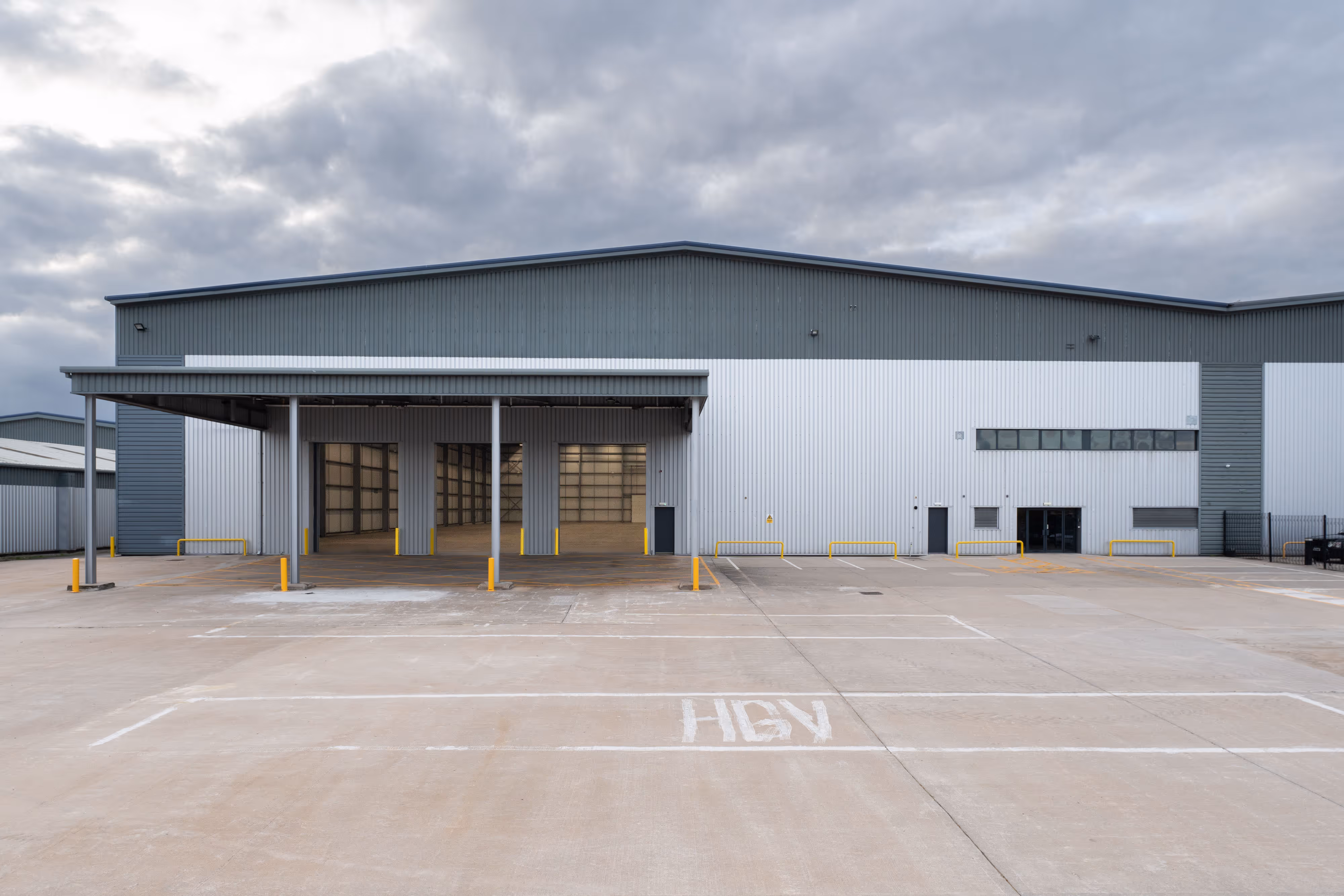 Large industrial warehouse with loading bays and empty concrete yard under a cloudy sky.