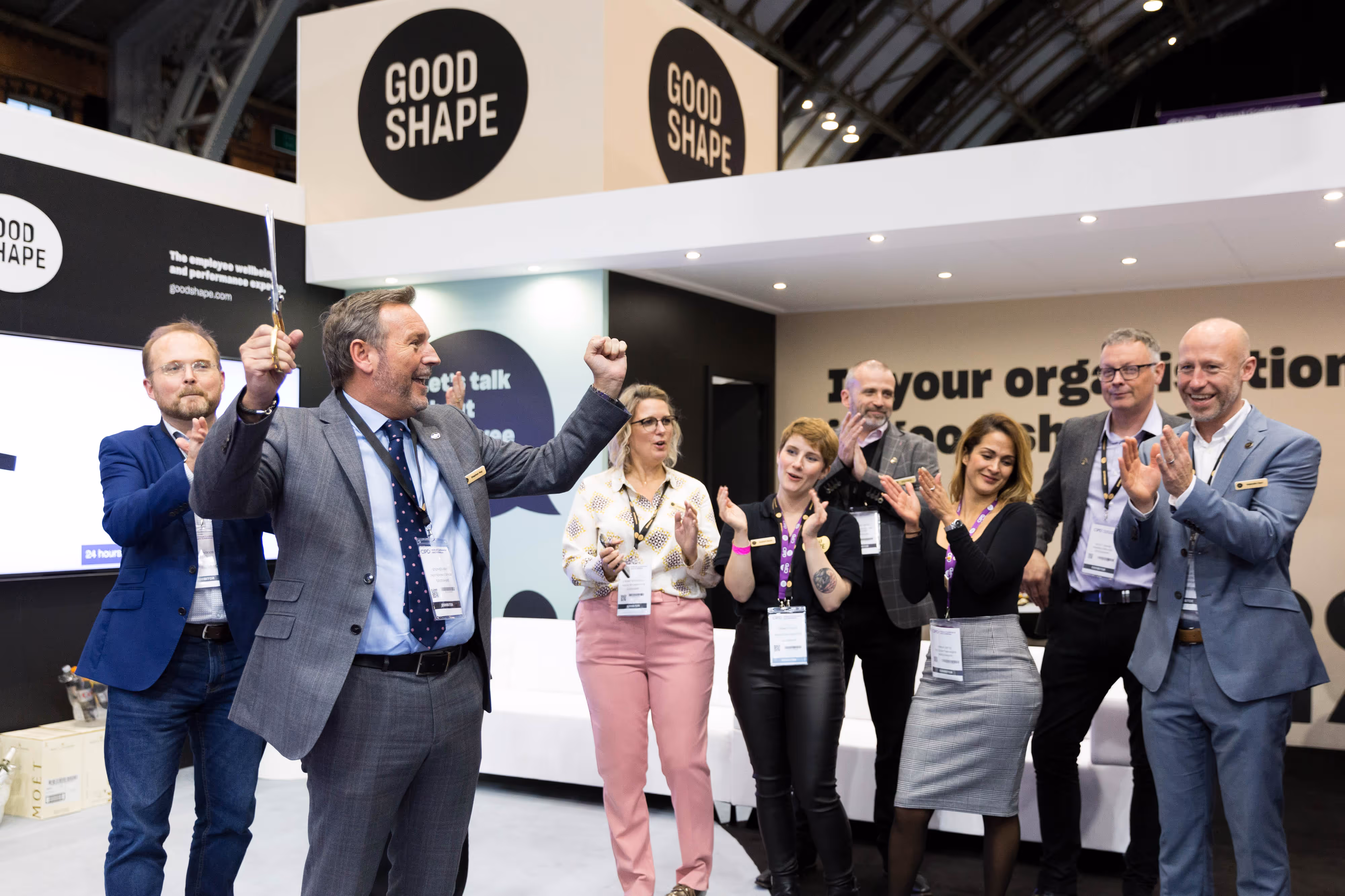 A group of people in business attire celebrate and clap near a stand labelled "GOOD SHAPE" at an event.