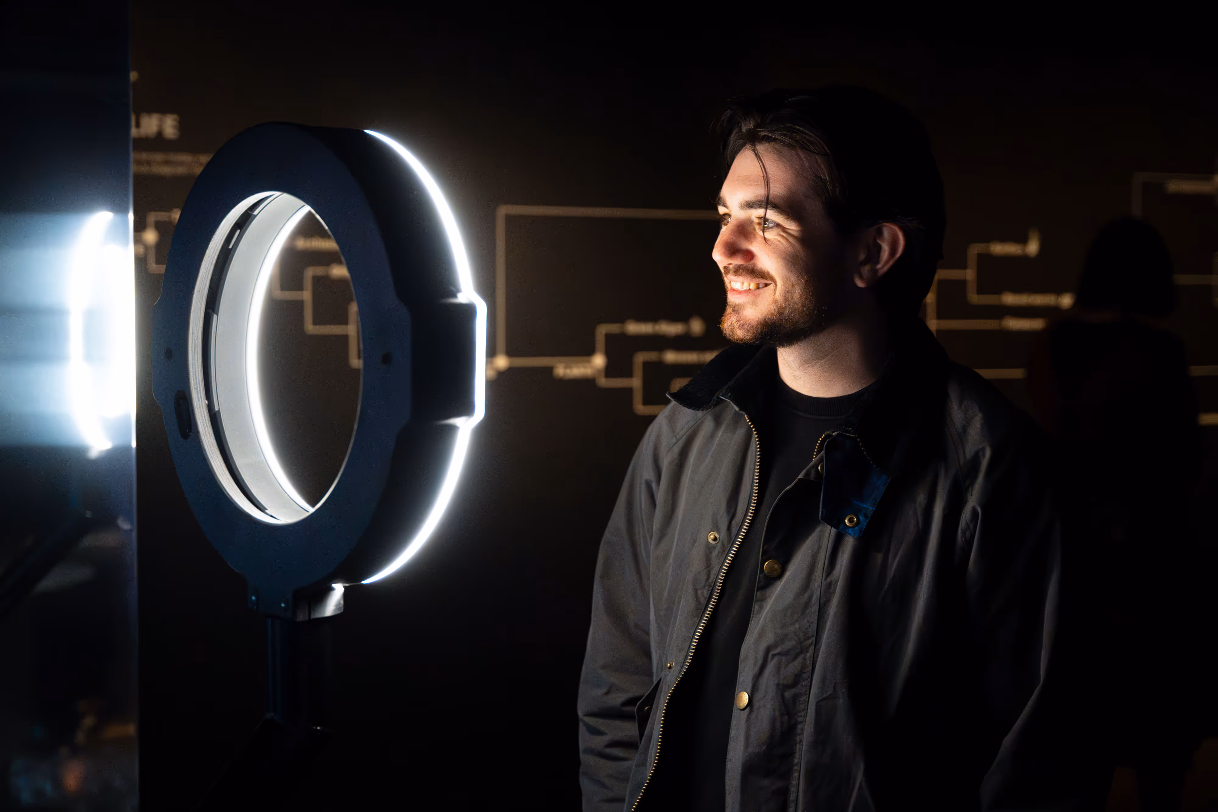 A man smiles while engaging with a ring light at an art exhibition in a dark room.