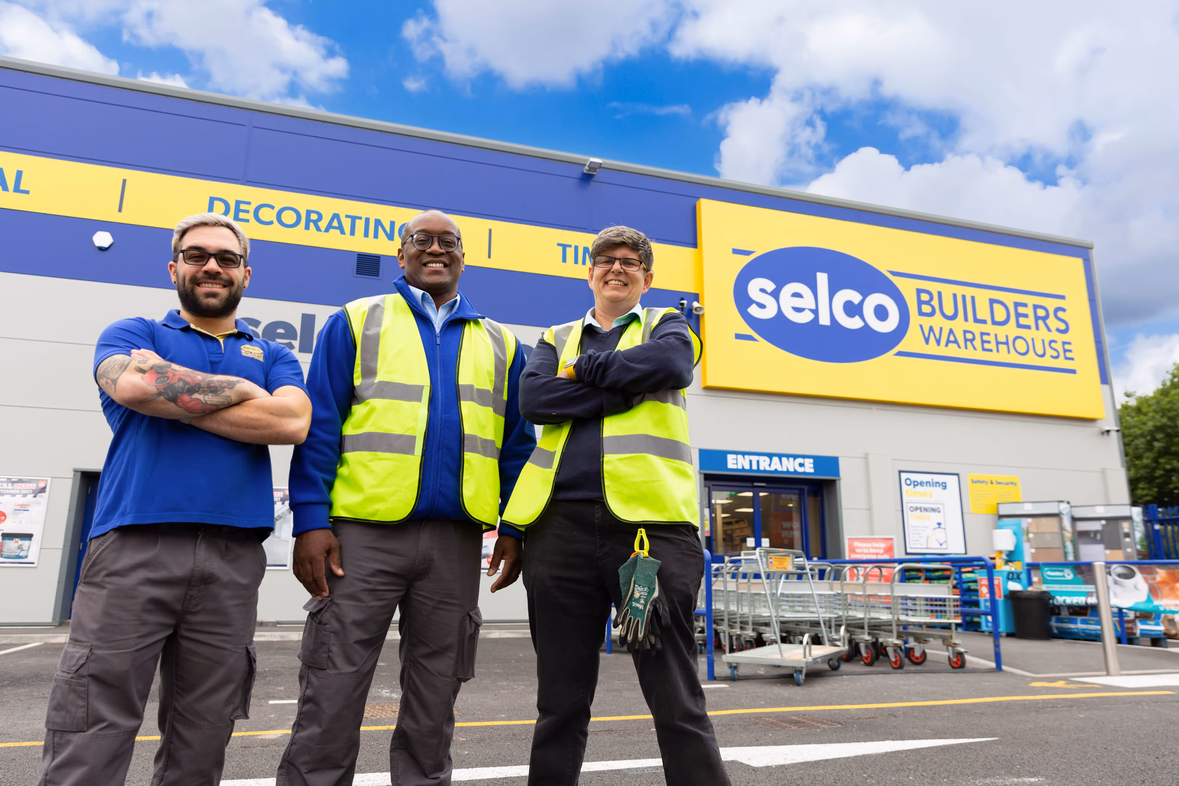 Three Selco Builders Warehouse employees stand smiling outside the store, two wearing yellow safety vests.