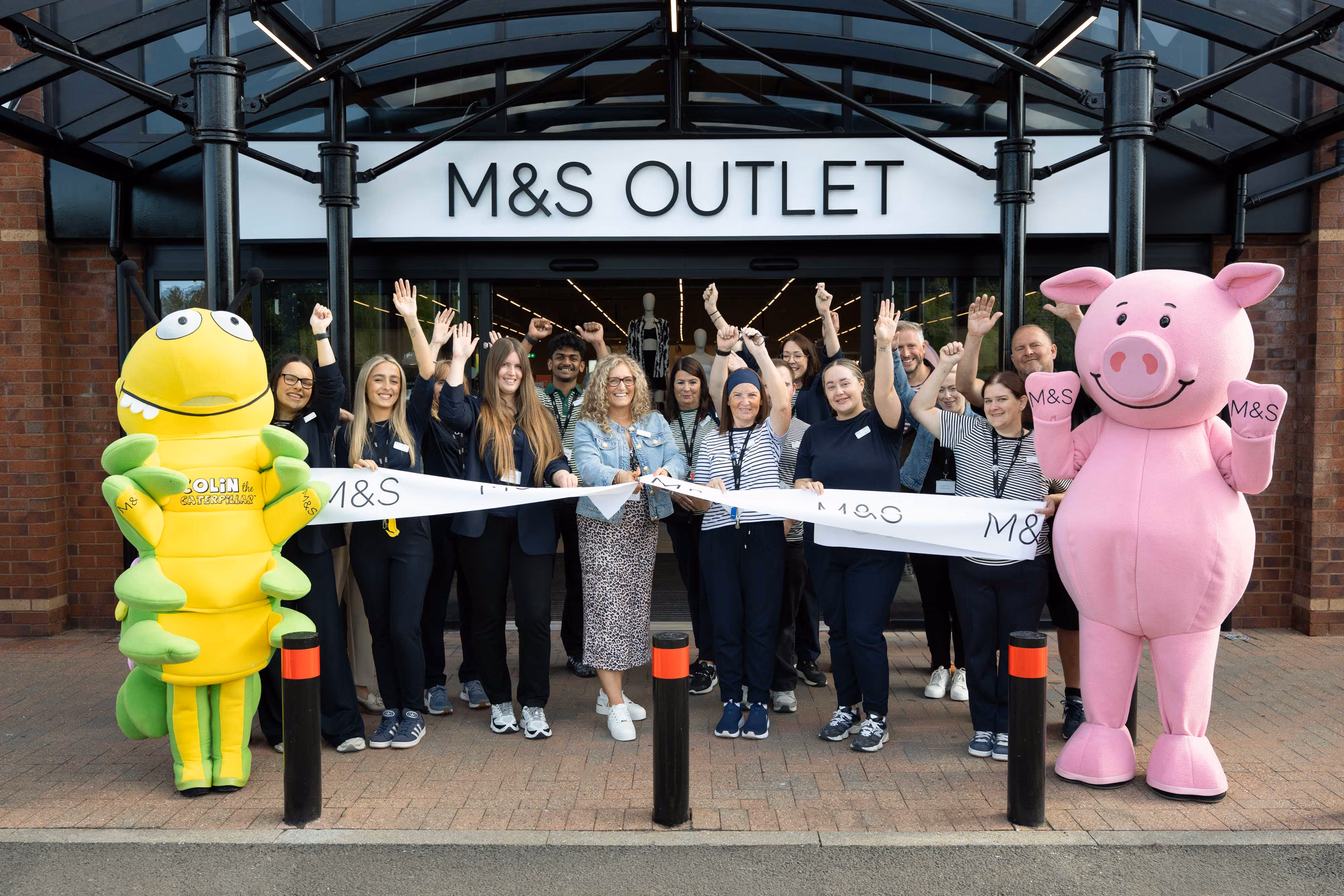 A group celebrates a ribbon cutting at an M&S Outlet, with Percy Pig and Colin the Caterpillar mascots.