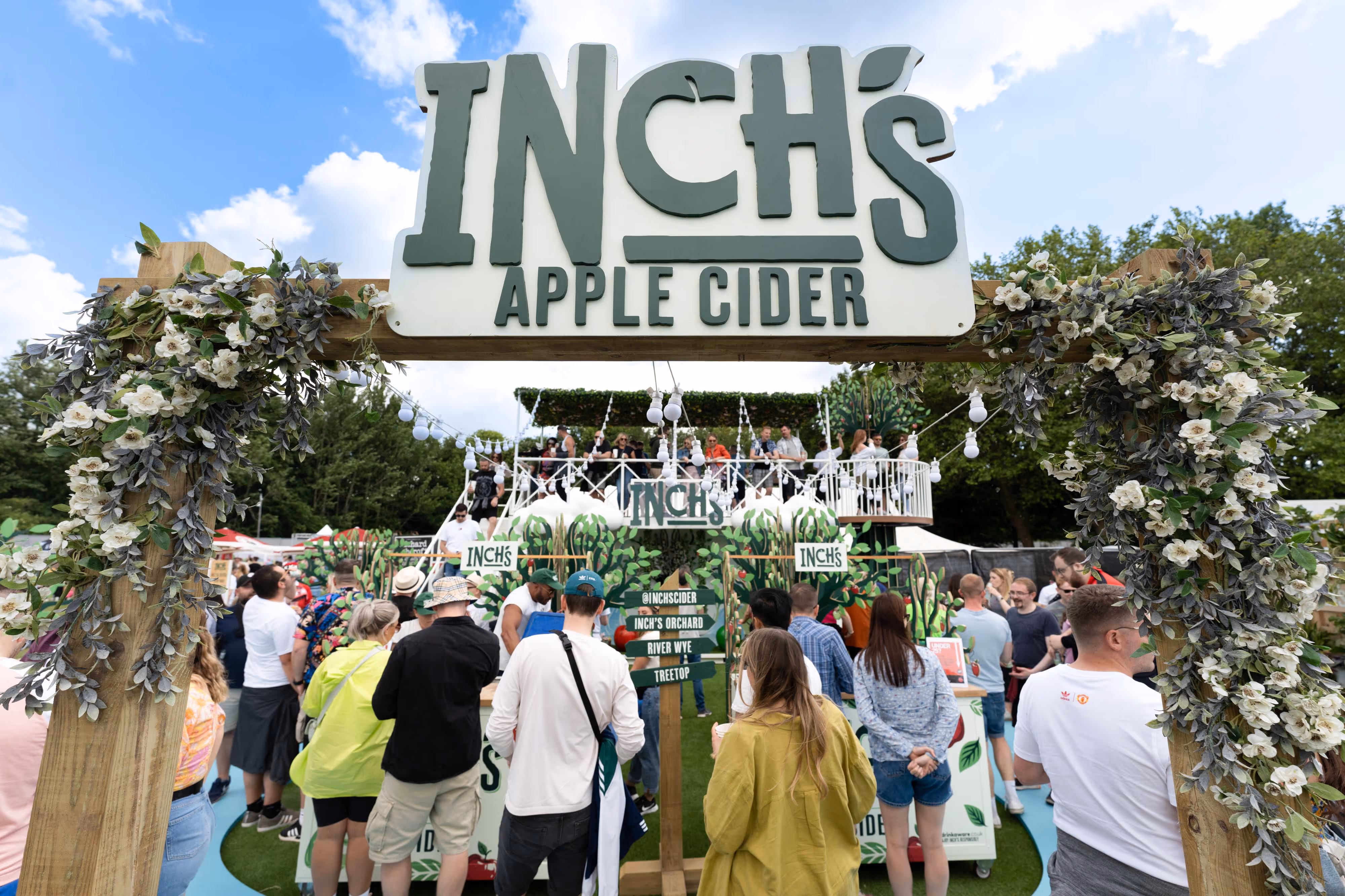 Crowd gathers at an outdoor event under a large "Inch's Apple Cider" sign decorated with greenery and flowers.