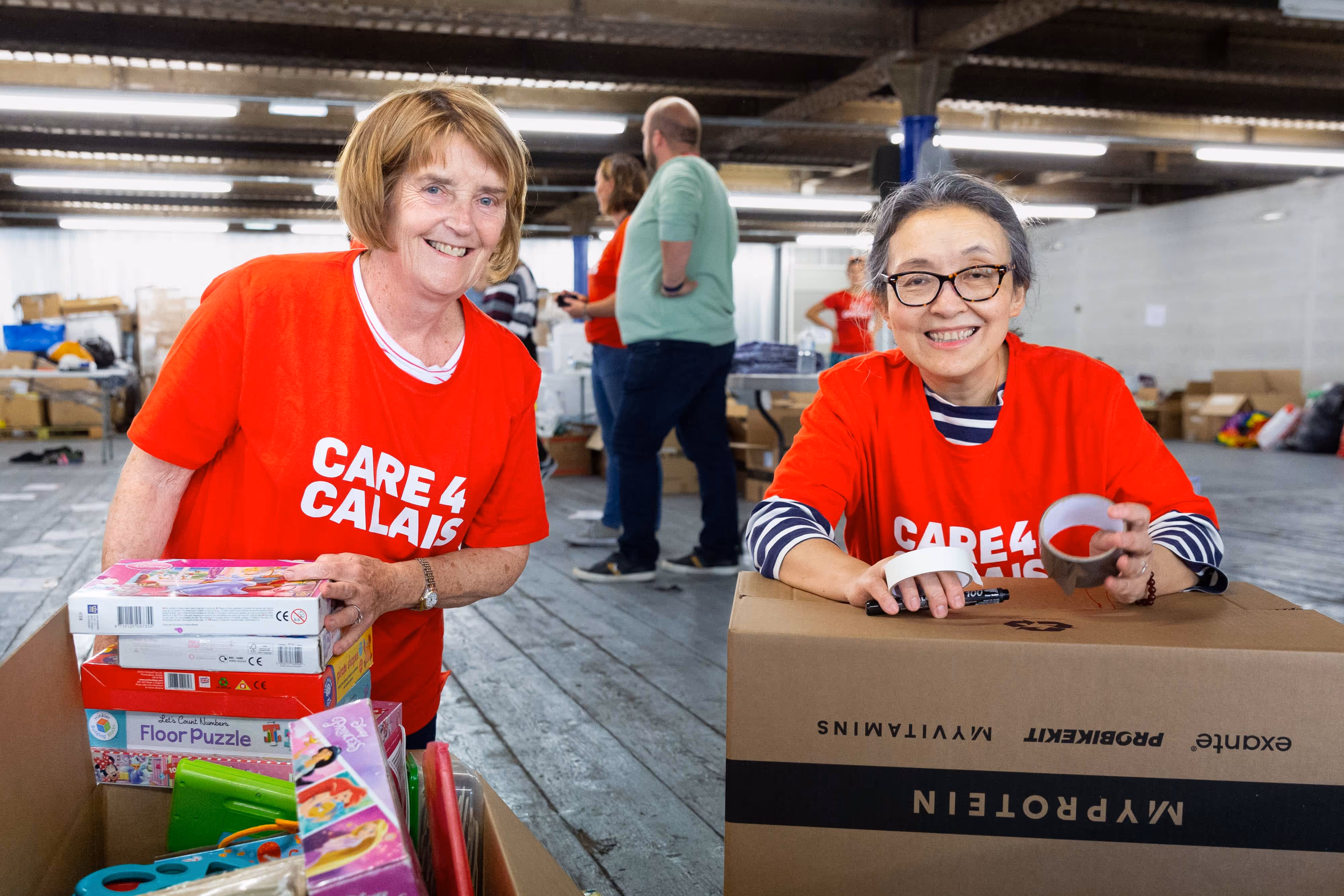 Two women in red "Care4Calais" shirts pack donated toys and supplies into boxes inside a warehouse.