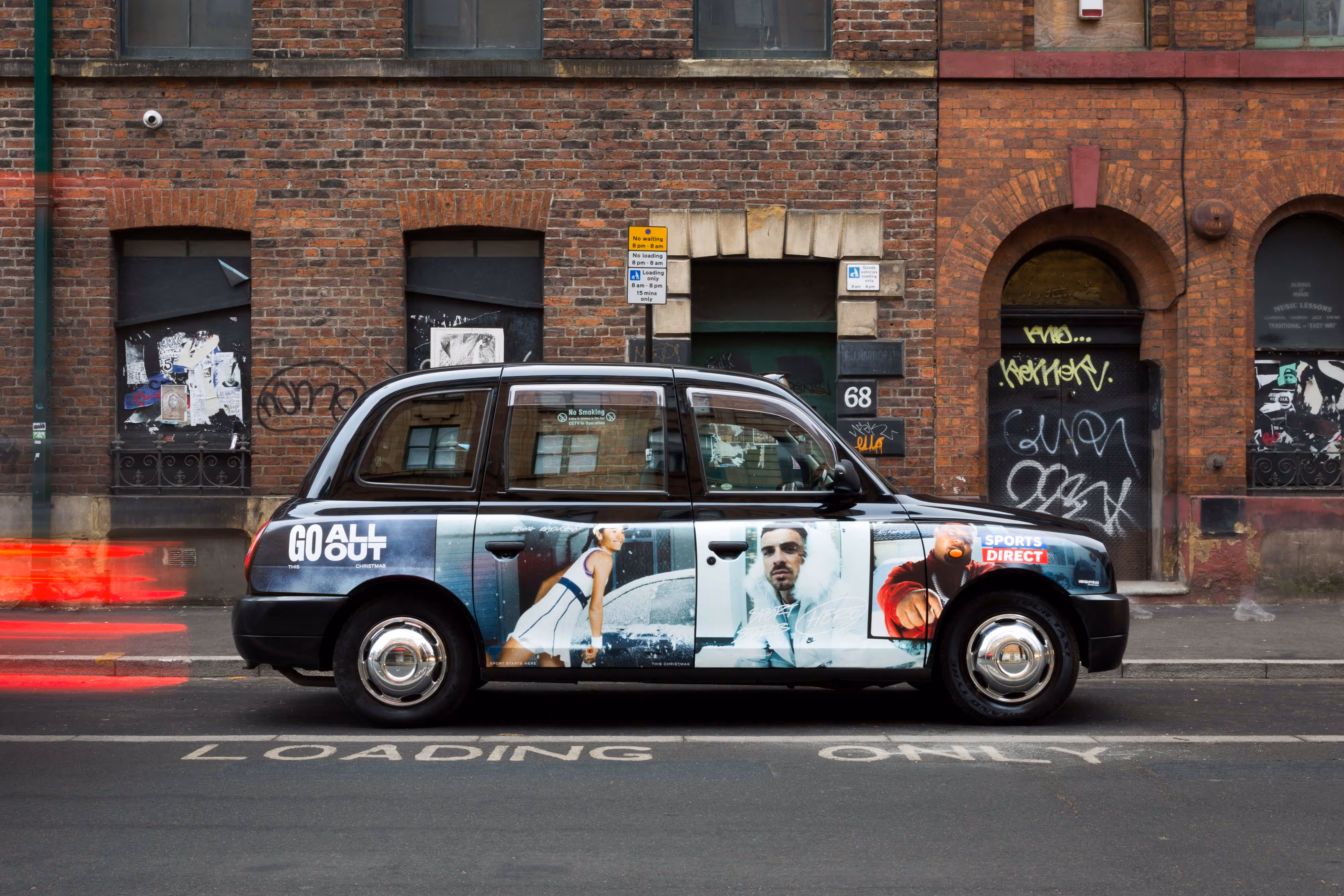 A black taxi with sports-themed adverts parked on a city street in front of a graffiti-covered brick building.