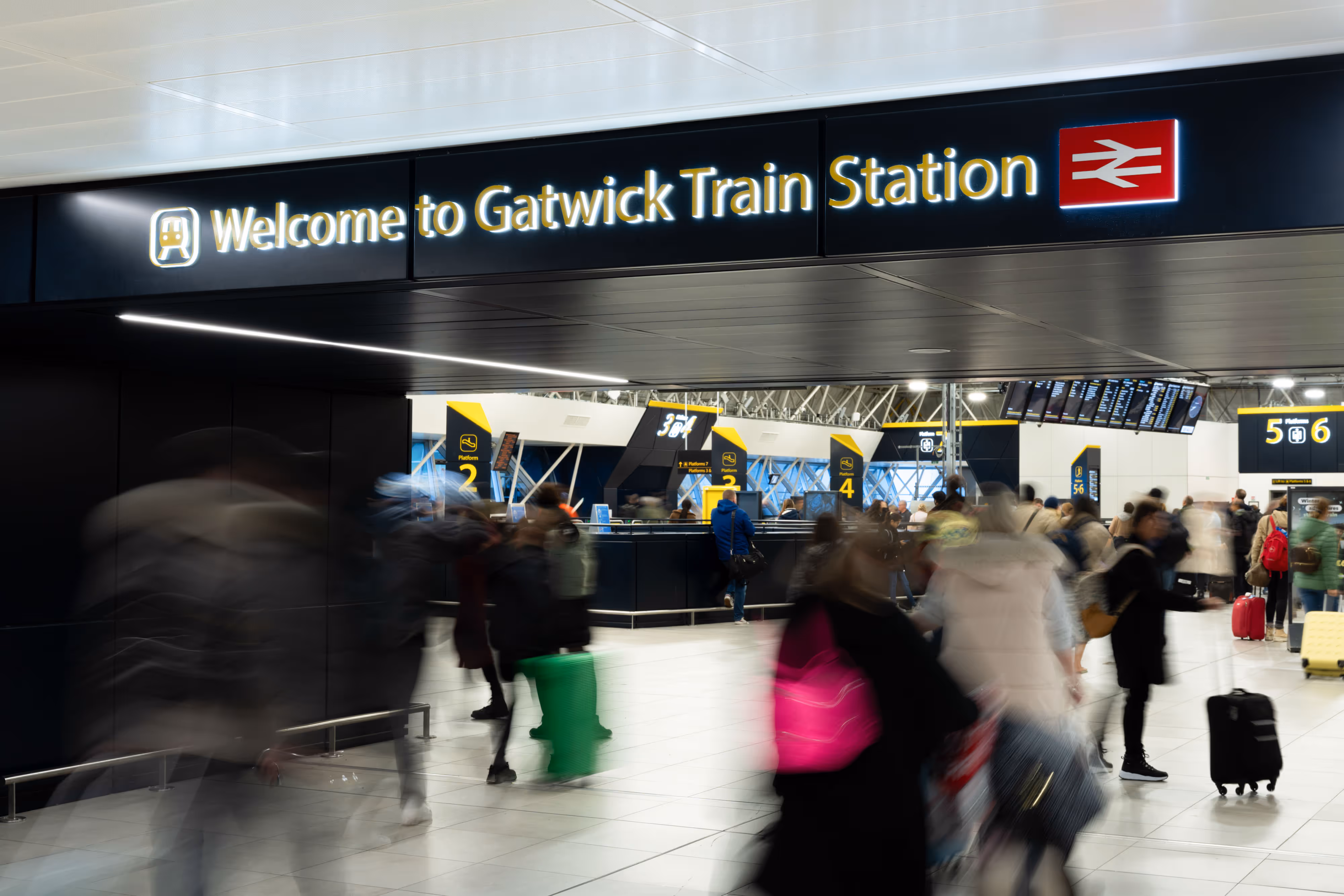 Busy scene at Gatwick Railway Station entrance with travellers and luggage beneath a bright welcome sign.