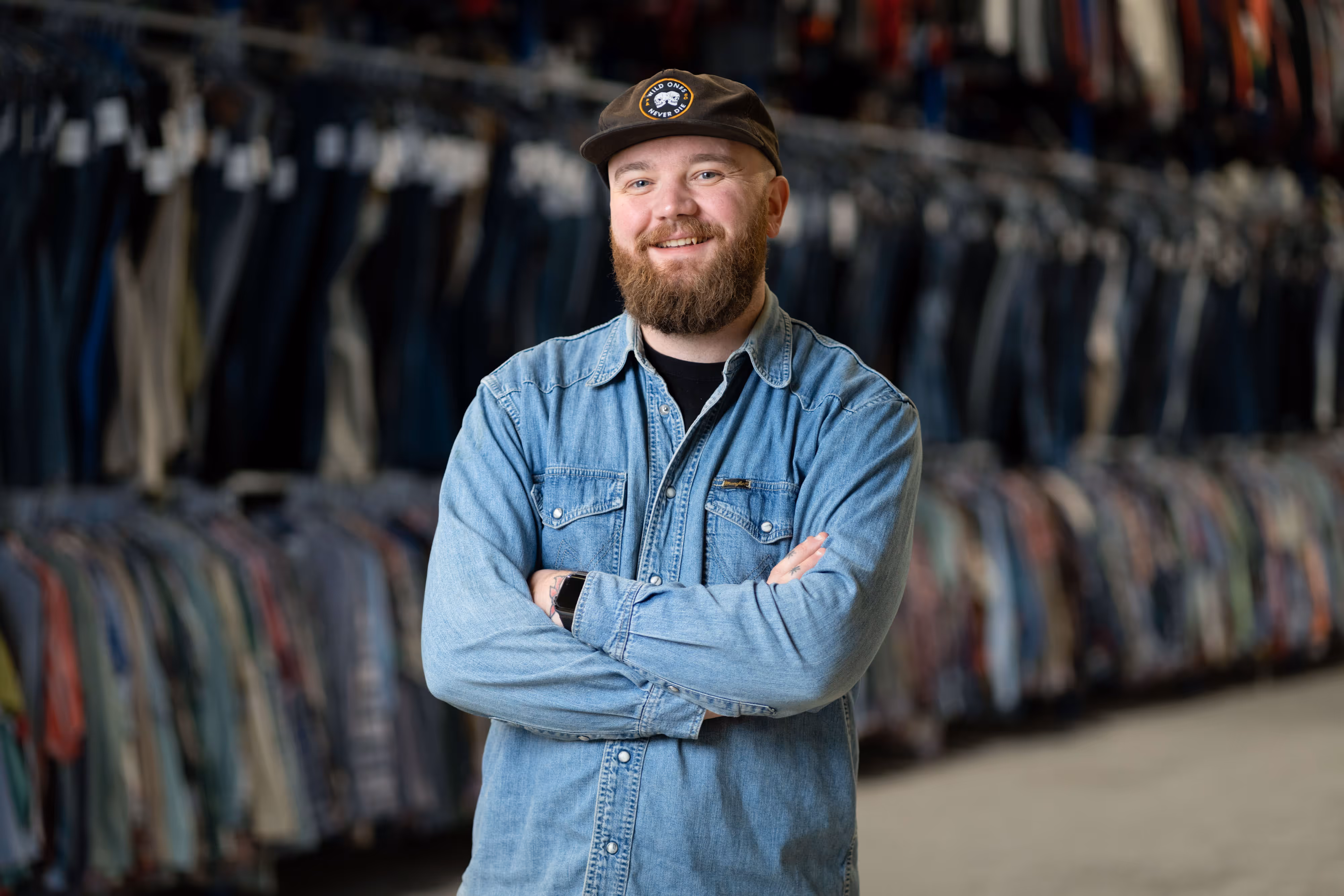 Bearded man in a denim shirt and cap smiles with arms folded, standing in front of rails of clothing.