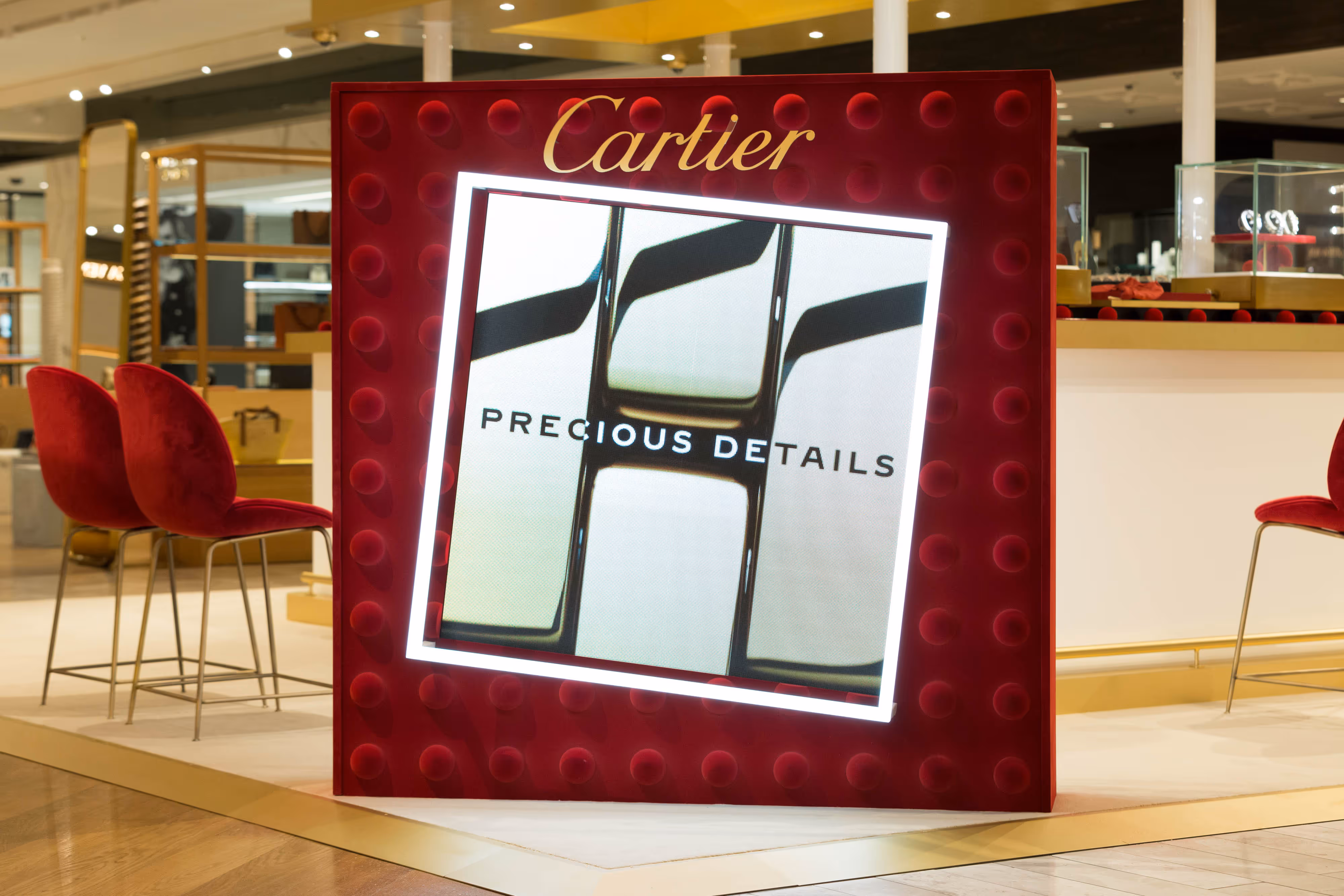 Red Cartier display sign with the words "Precious Details" in a lit frame, near red chairs in a shopping centre setting.