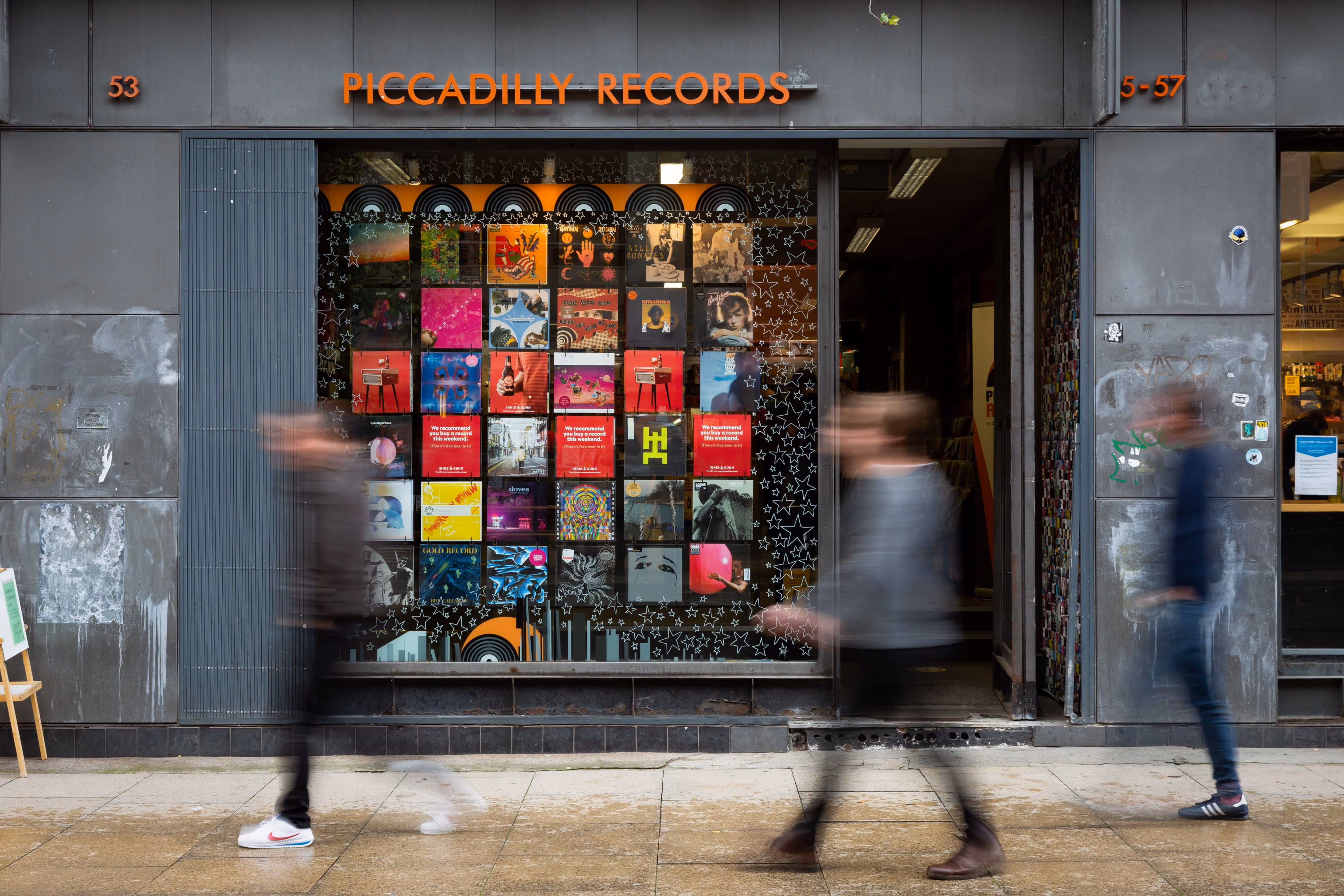 Three people walk past Piccadilly Records shop with colourful album covers displayed in the window.