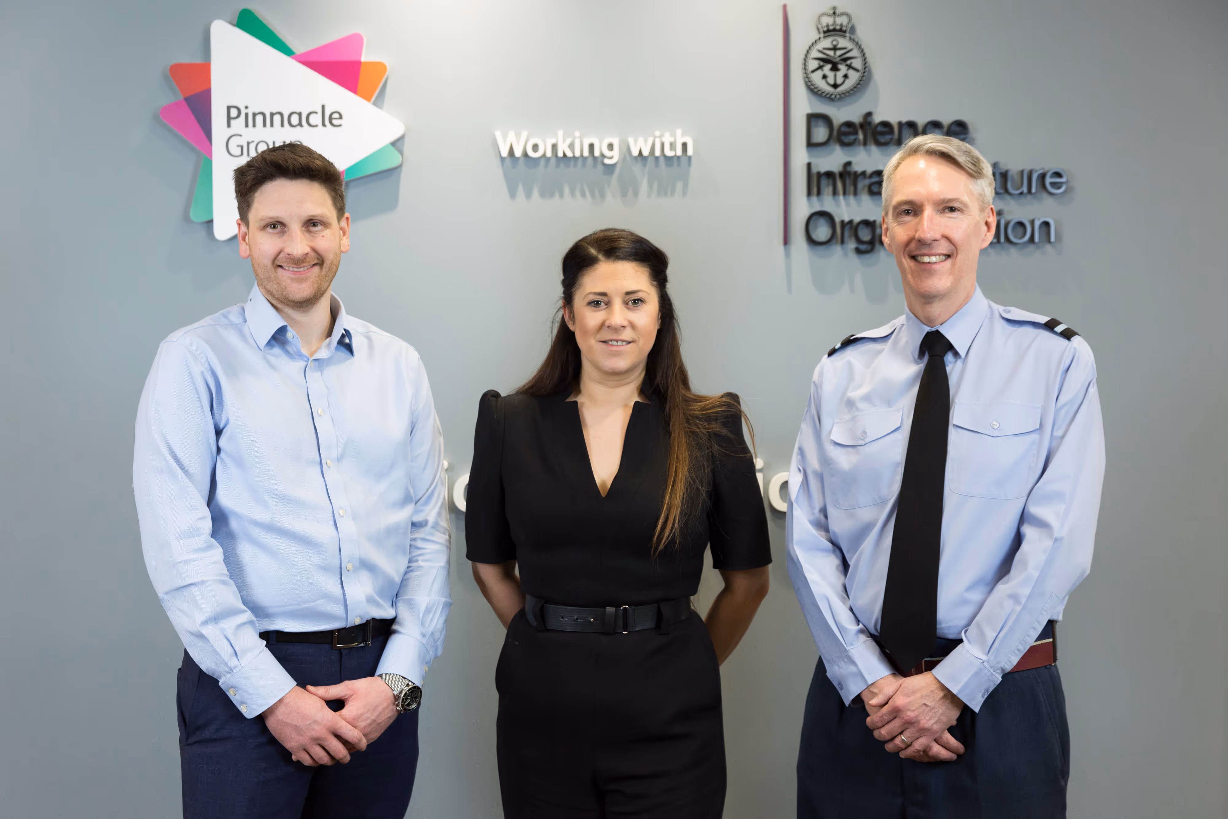 Three people stand smiling in front of Pinnacle Group and Defence Infrastructure Organisation logos on a grey wall.