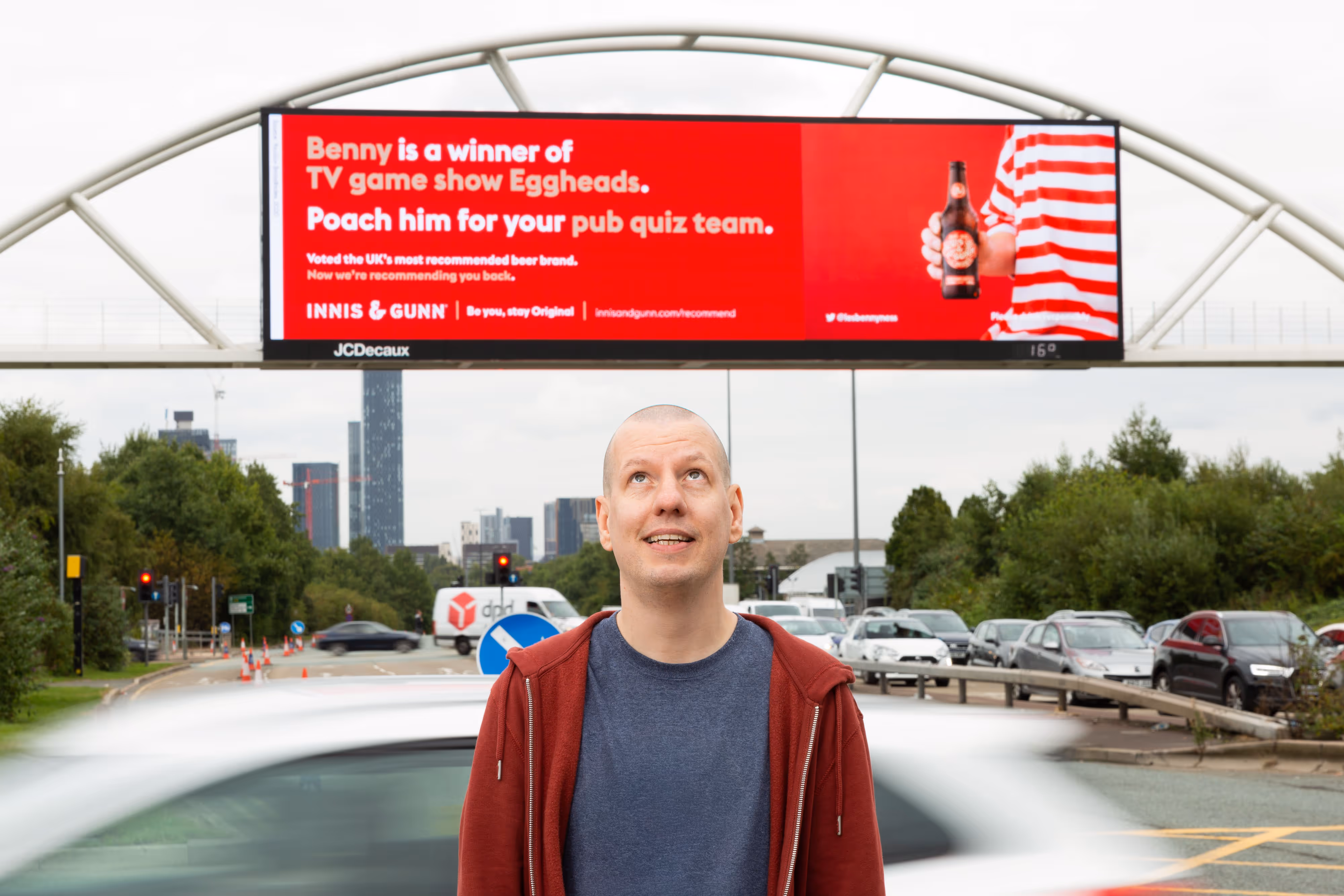 A man standing in front of a red billboard for a Scottish beer brand which features him.