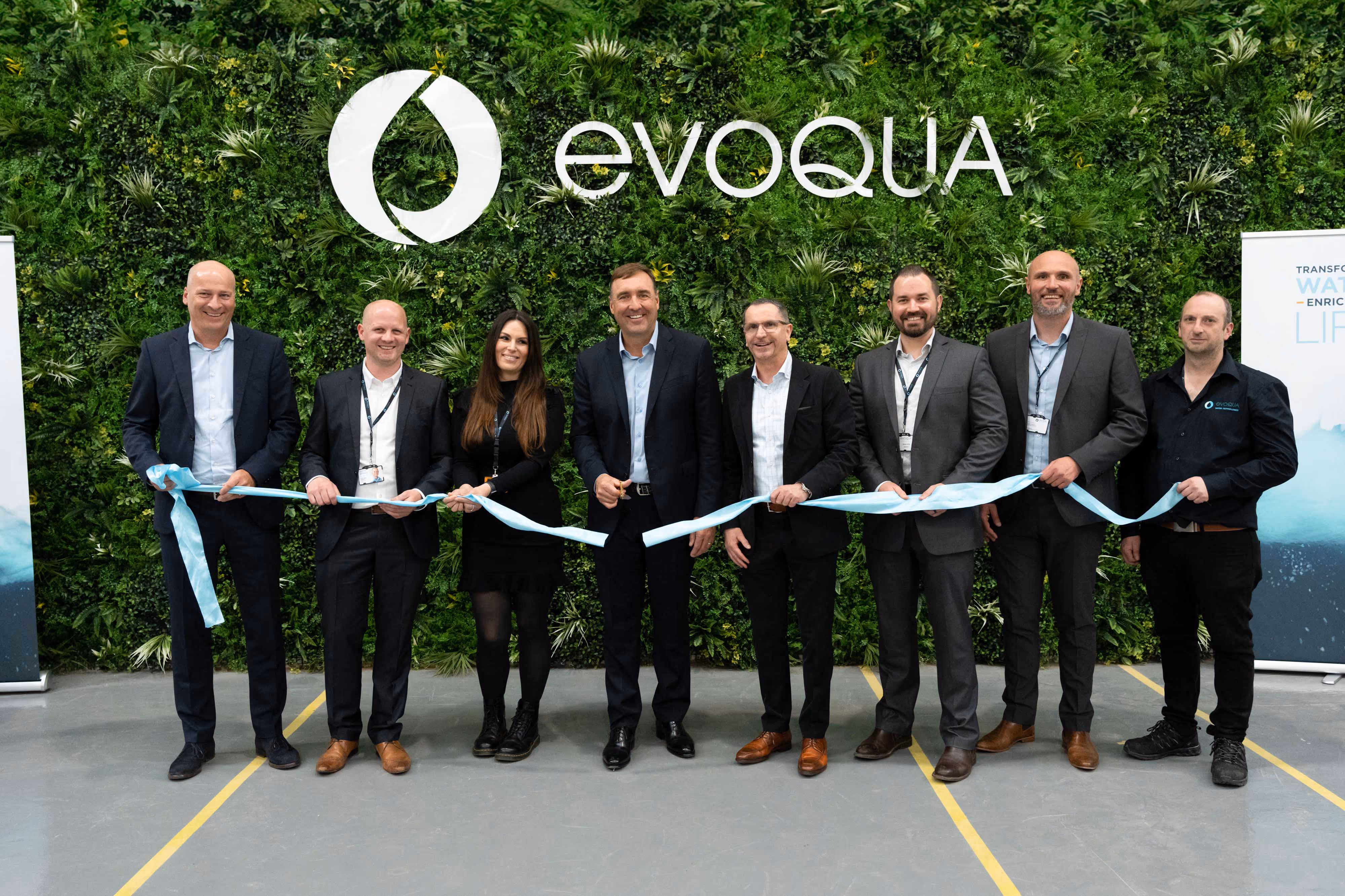 Eight people in business attire cut a blue ribbon in front of a green wall with the Evoqua logo.