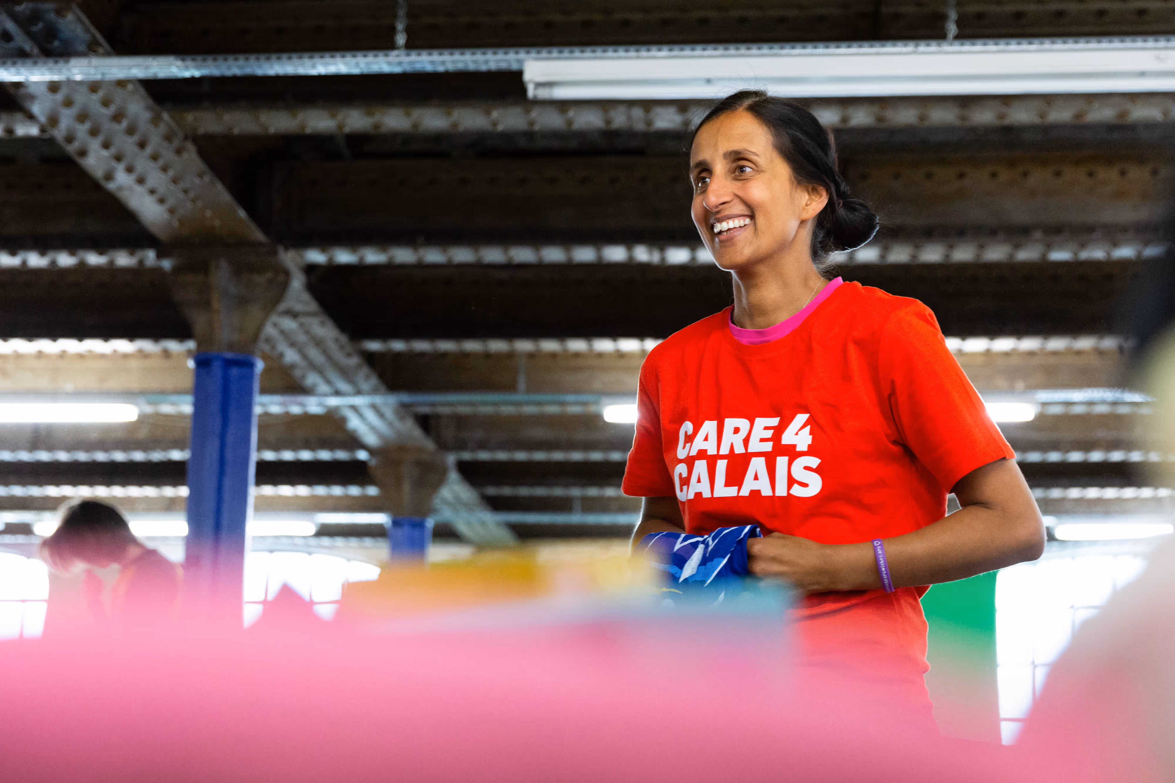 Smiling woman wearing a red "Care 4 Calais" t-shirt stands indoors, holding blue fabric.