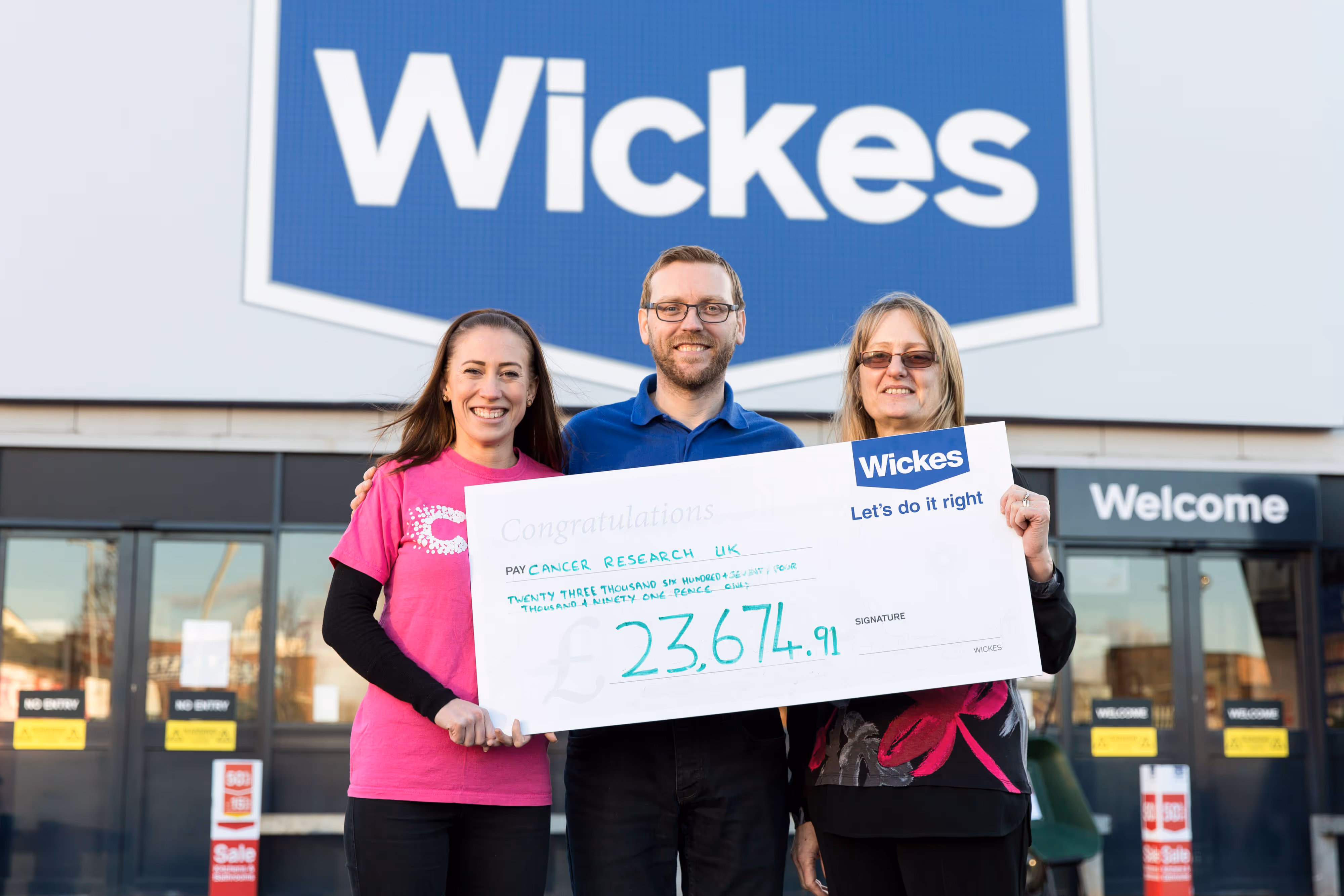 Three people smiling and holding a large cheque for outside a Wickes store, supporting Cancer Research UK.