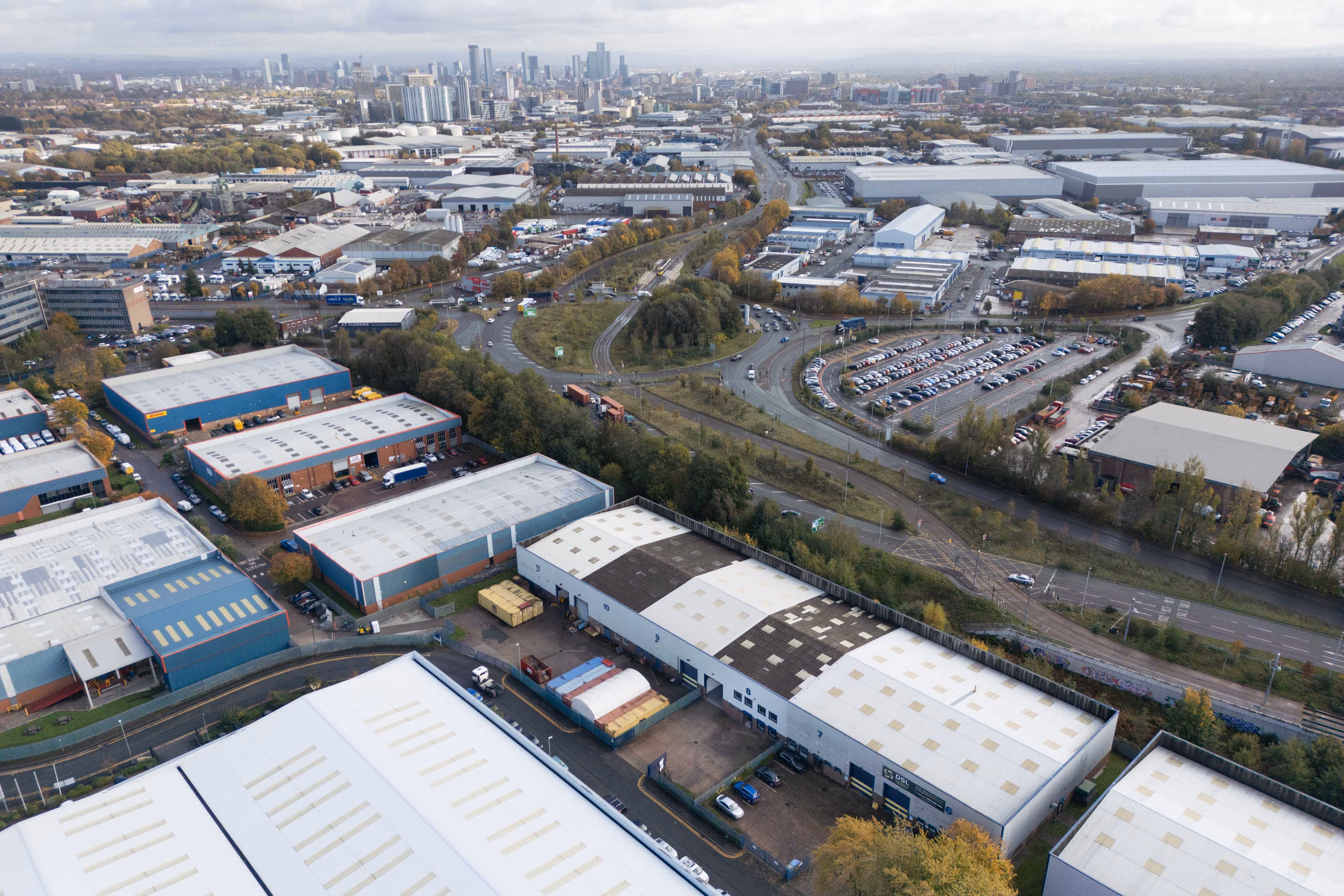 Aerial view of industrial buildings, roads, and car parks with a city skyline in the background.