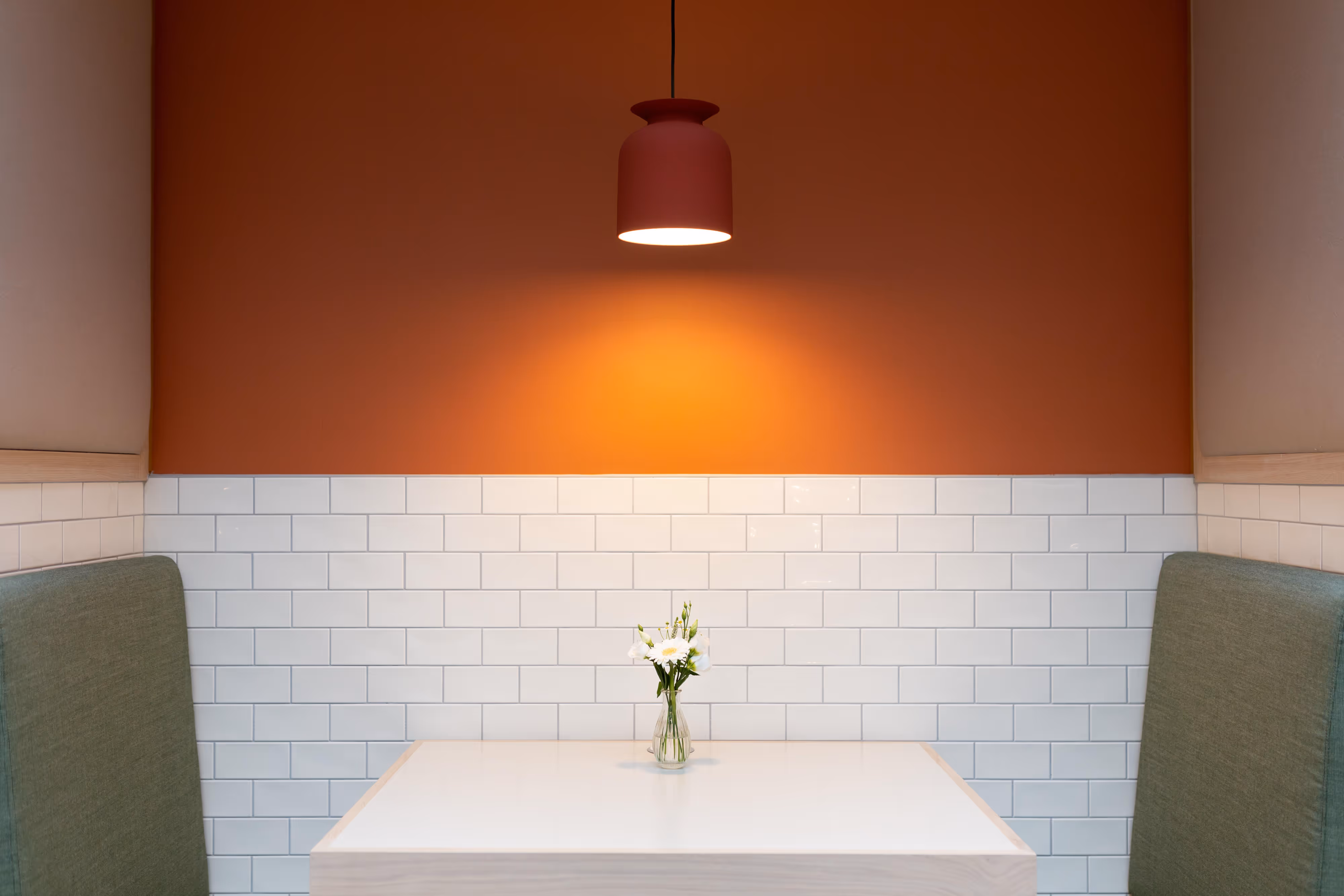 Small table with a flower vase in a booth, lit by a hanging lamp, with orange and white tiled walls.