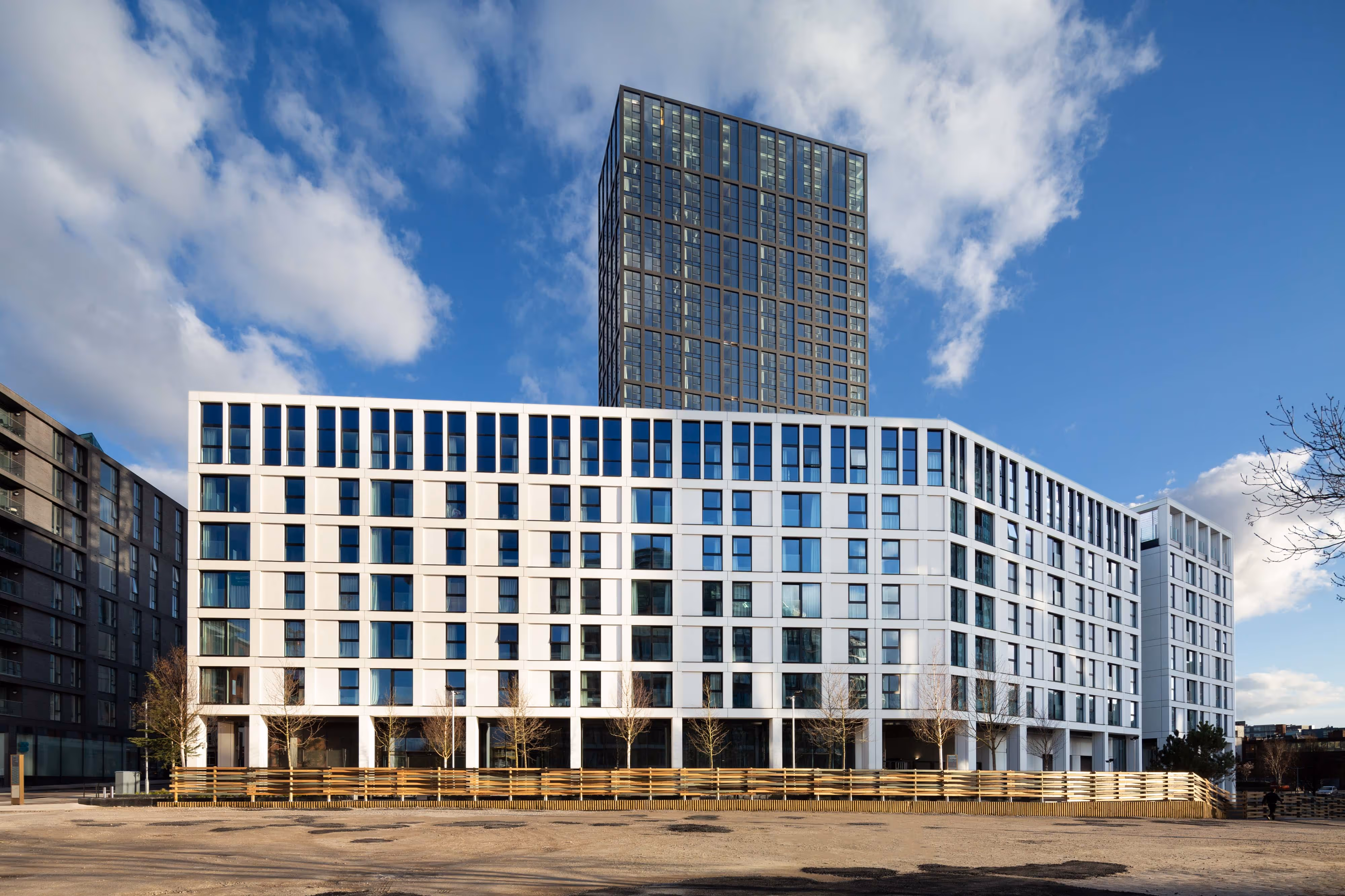 Modern multi-storey building with white facade and a tall glass tower in the background under a partly cloudy sky.