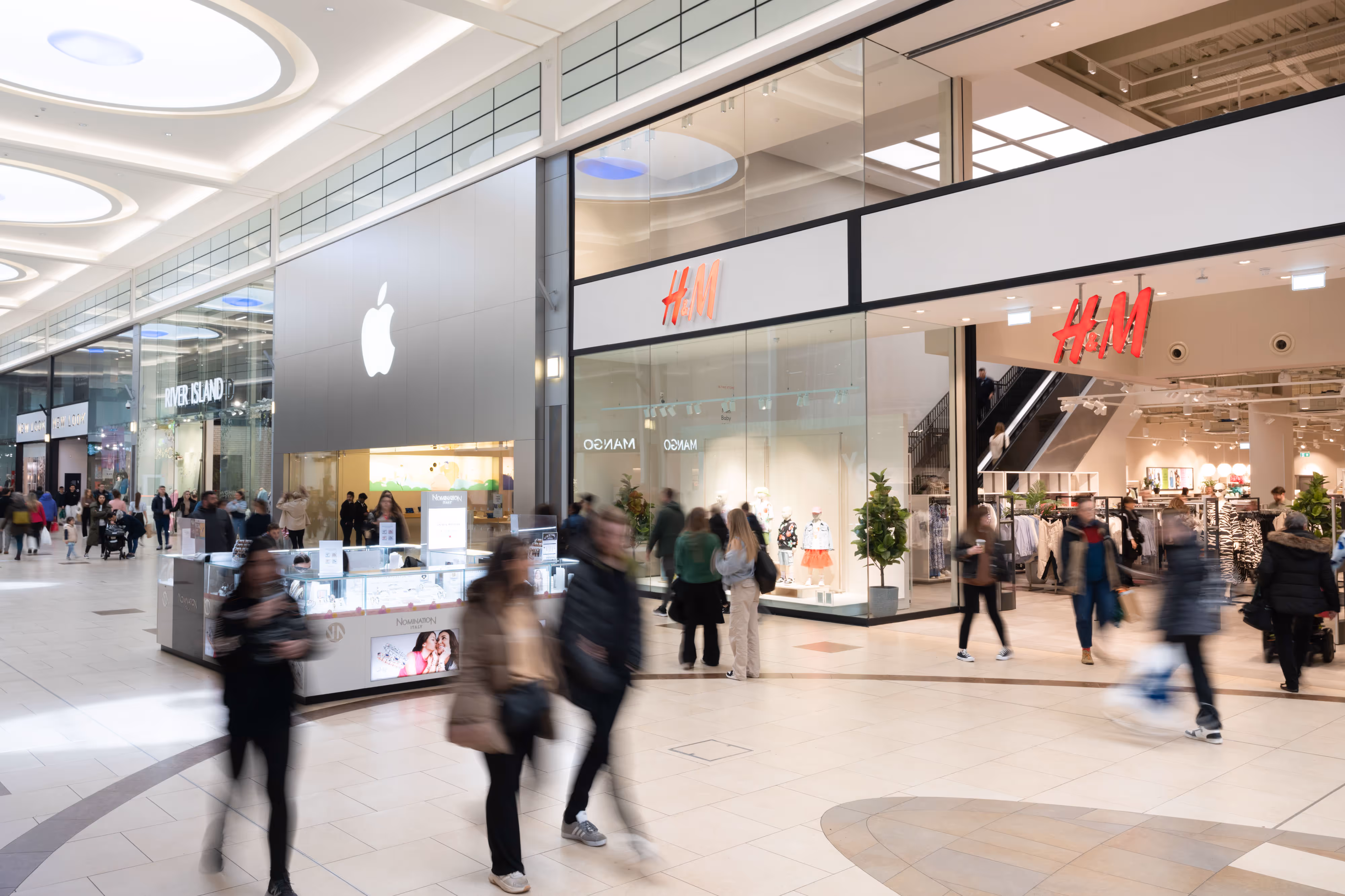 Shoppers walk past Apple and H&M stores inside a bright, modern shopping centre.