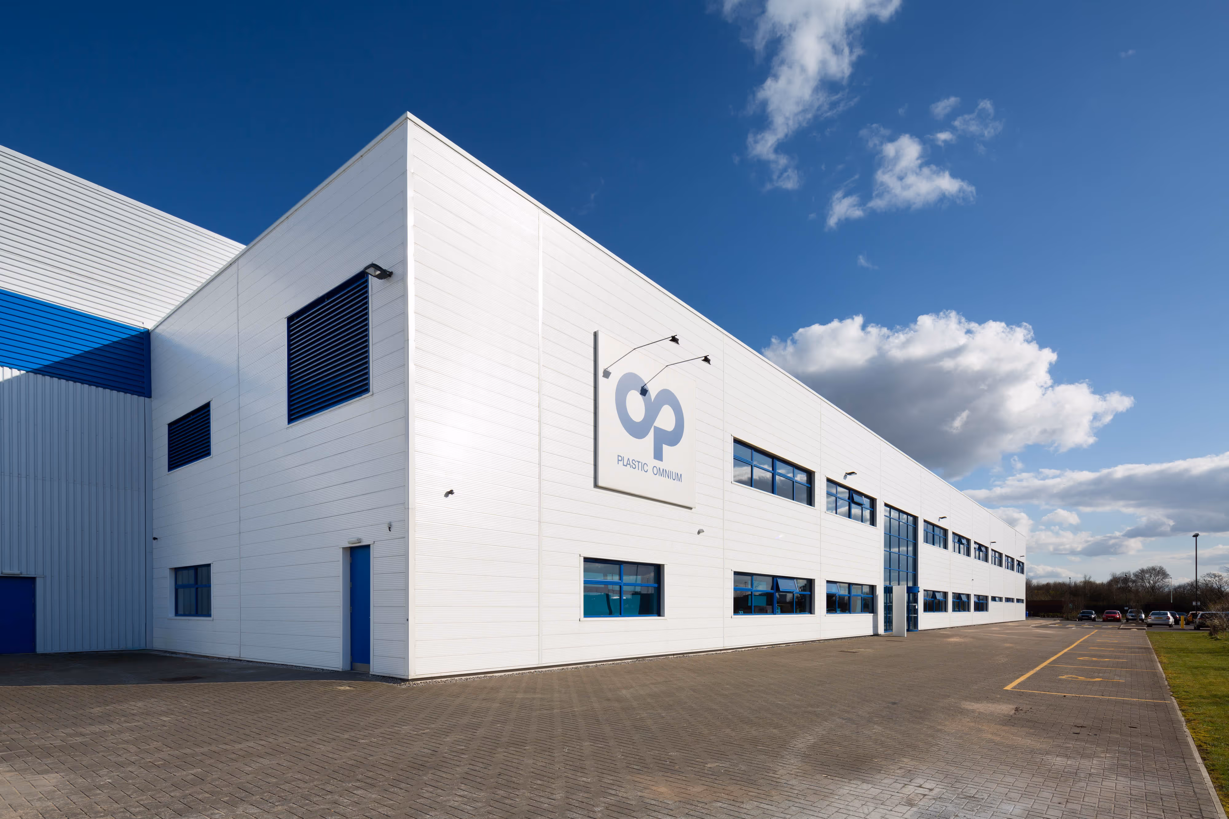 Large white industrial building with blue accents, tall windows, and a logo on the side under a blue sky near Warrington.