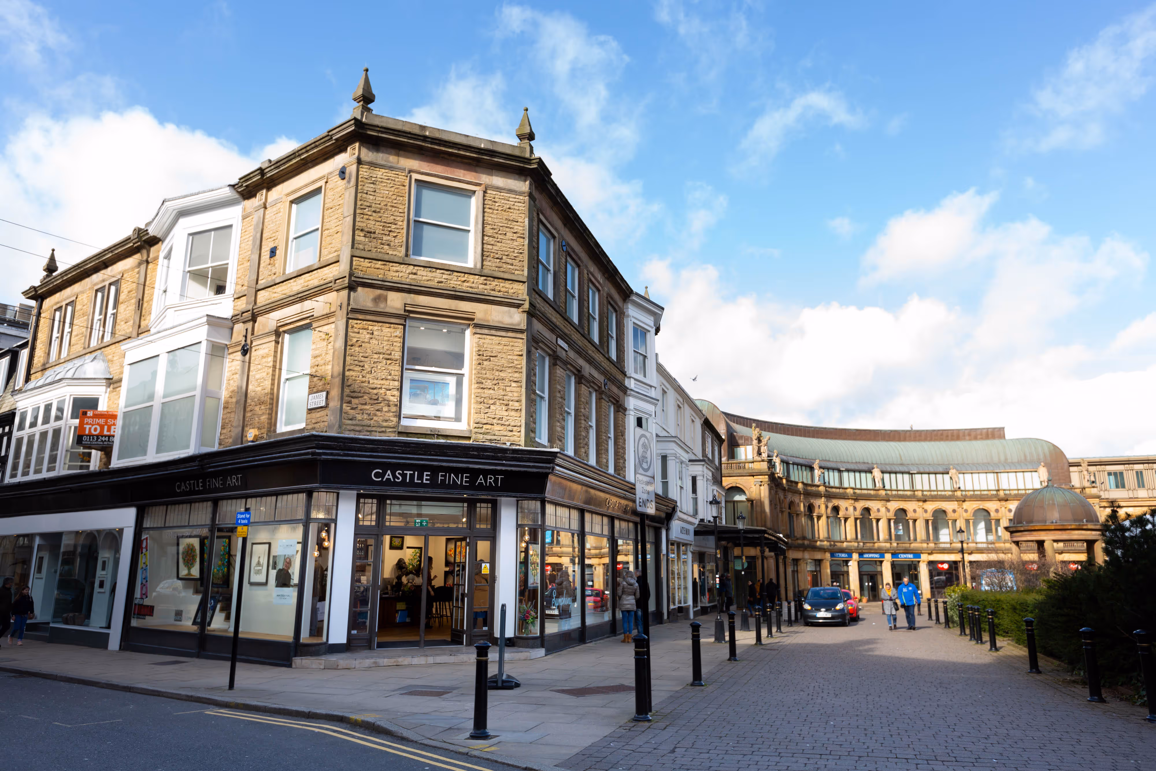 A street corner in Harrogate with a Castle Fine Art gallery, shops, and people walking under a partly cloudy sky.
