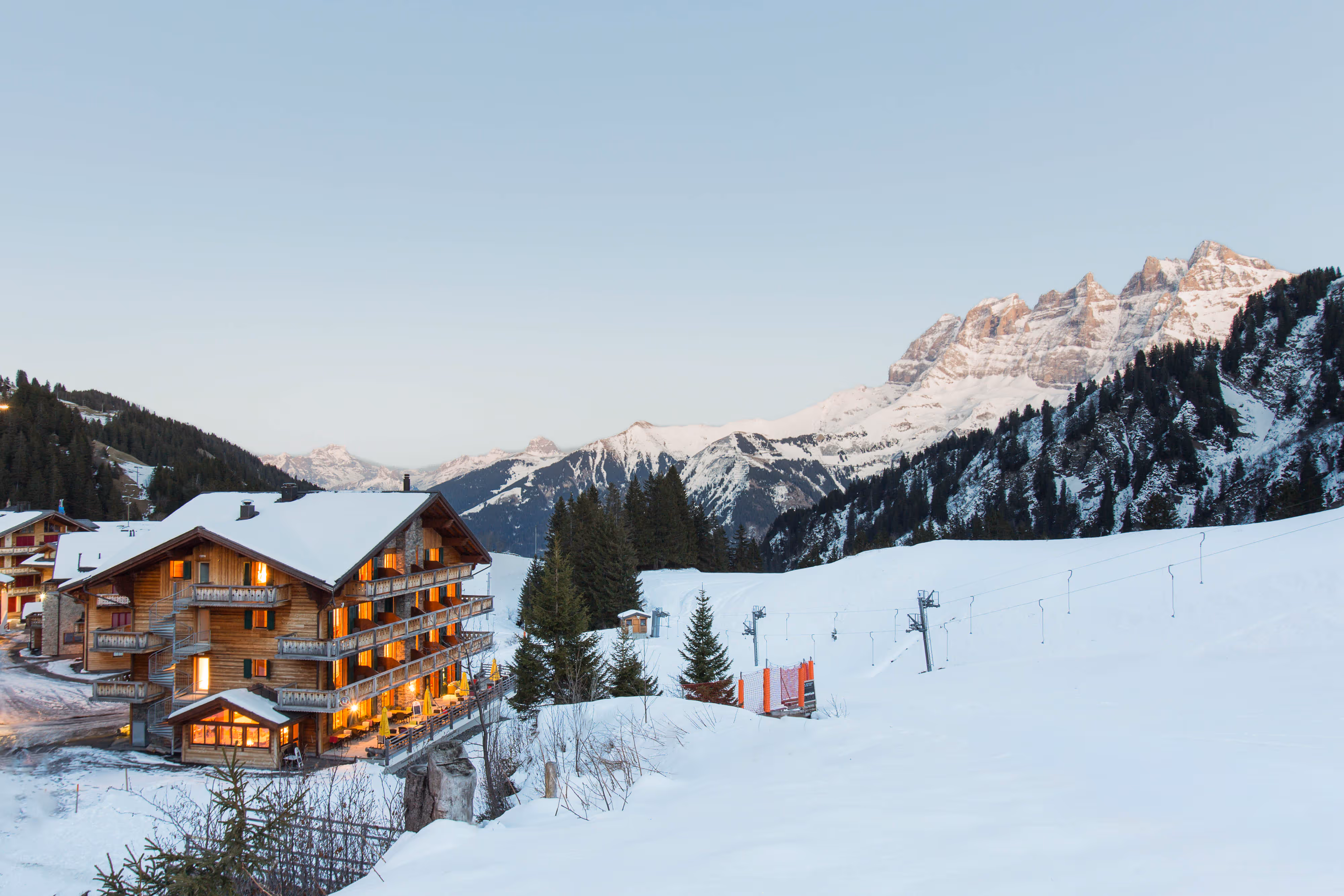 A wooden chalet with glowing lights sits among snowy mountains and trees at sundown.