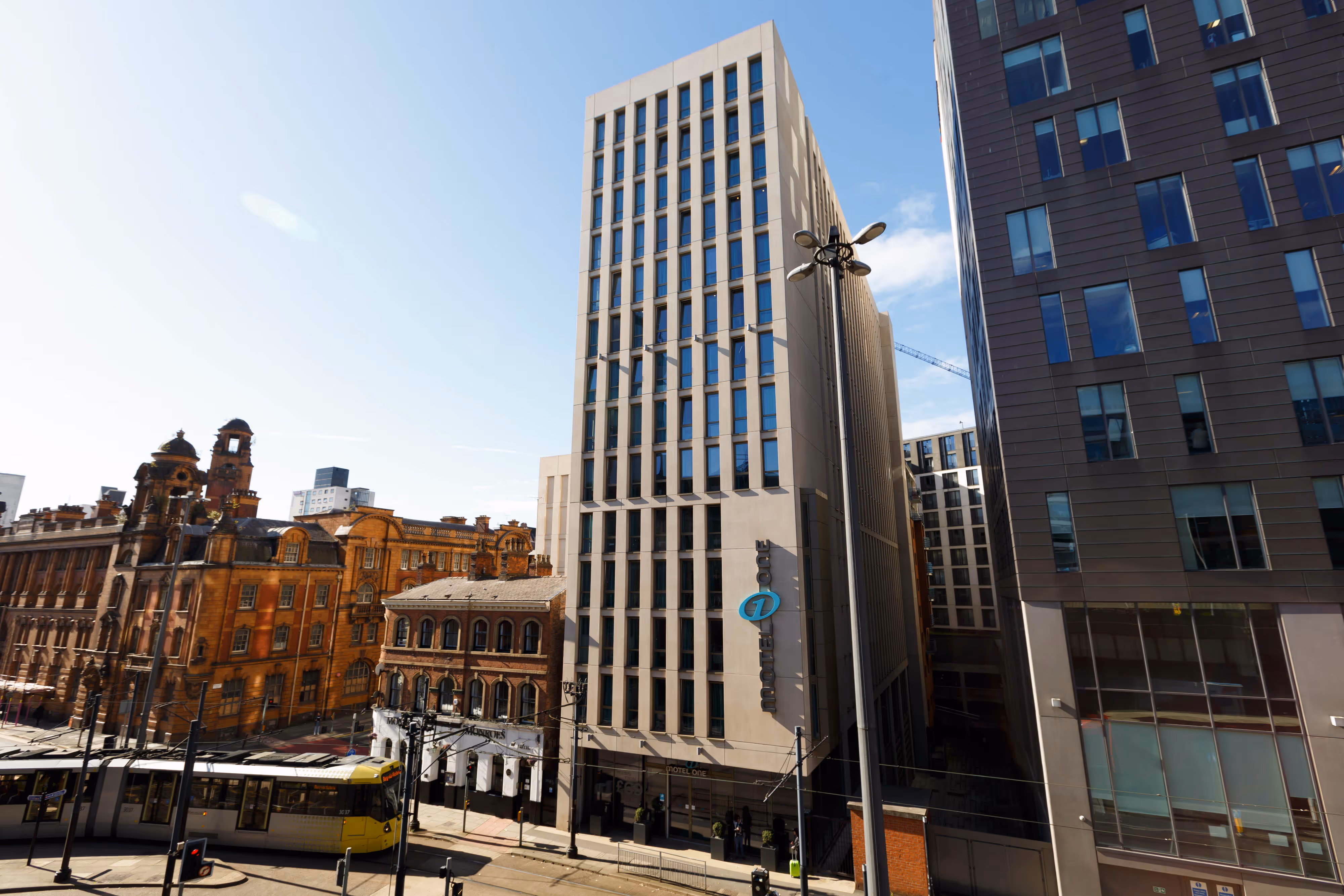 A modern high-rise building stands beside historic architecture on a sunny city street with trams and pedestrians visible.