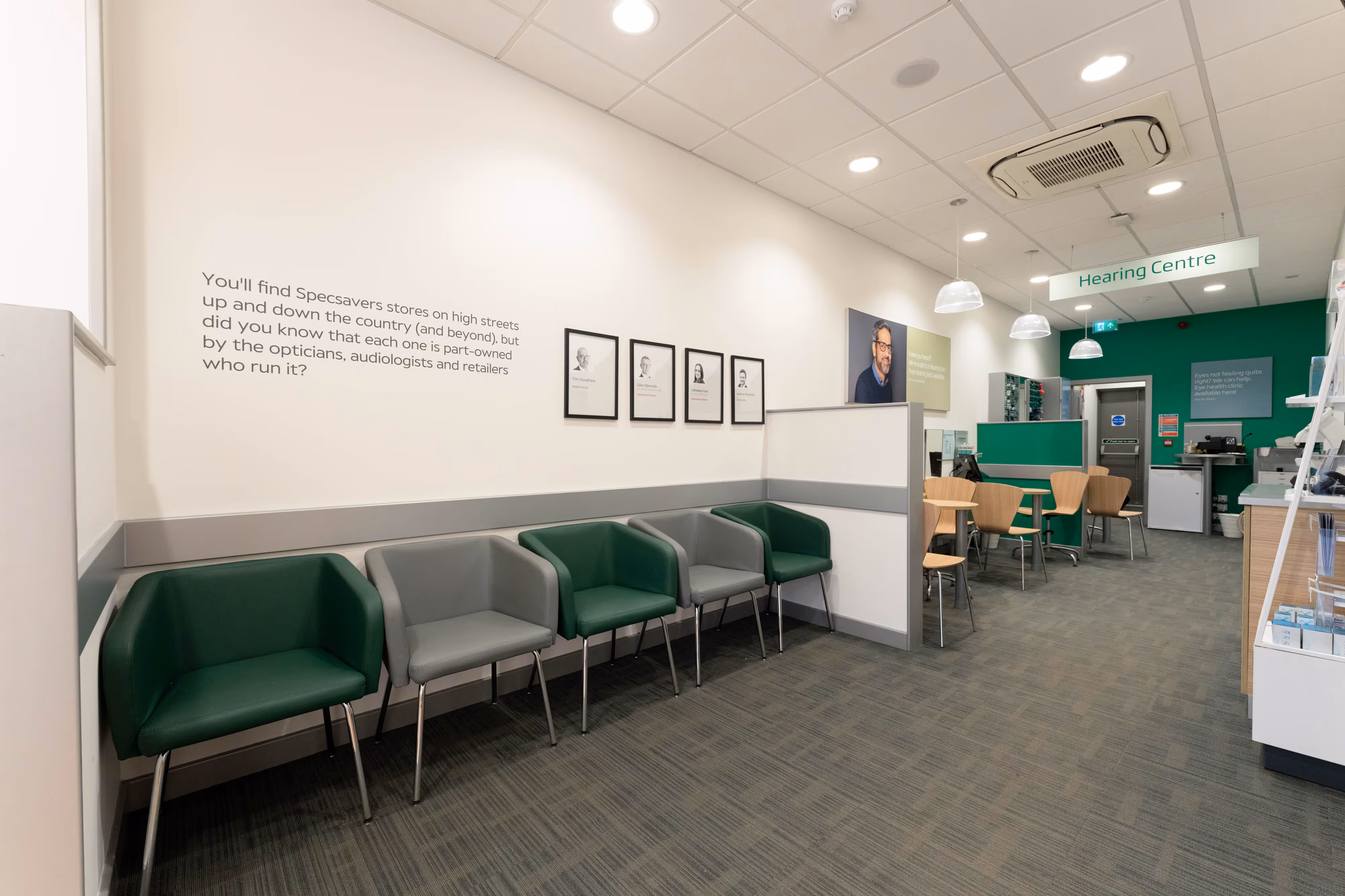 An empty optician's waiting area with green and grey chairs, wall portraits, and a sign reading "Hearing Centre".