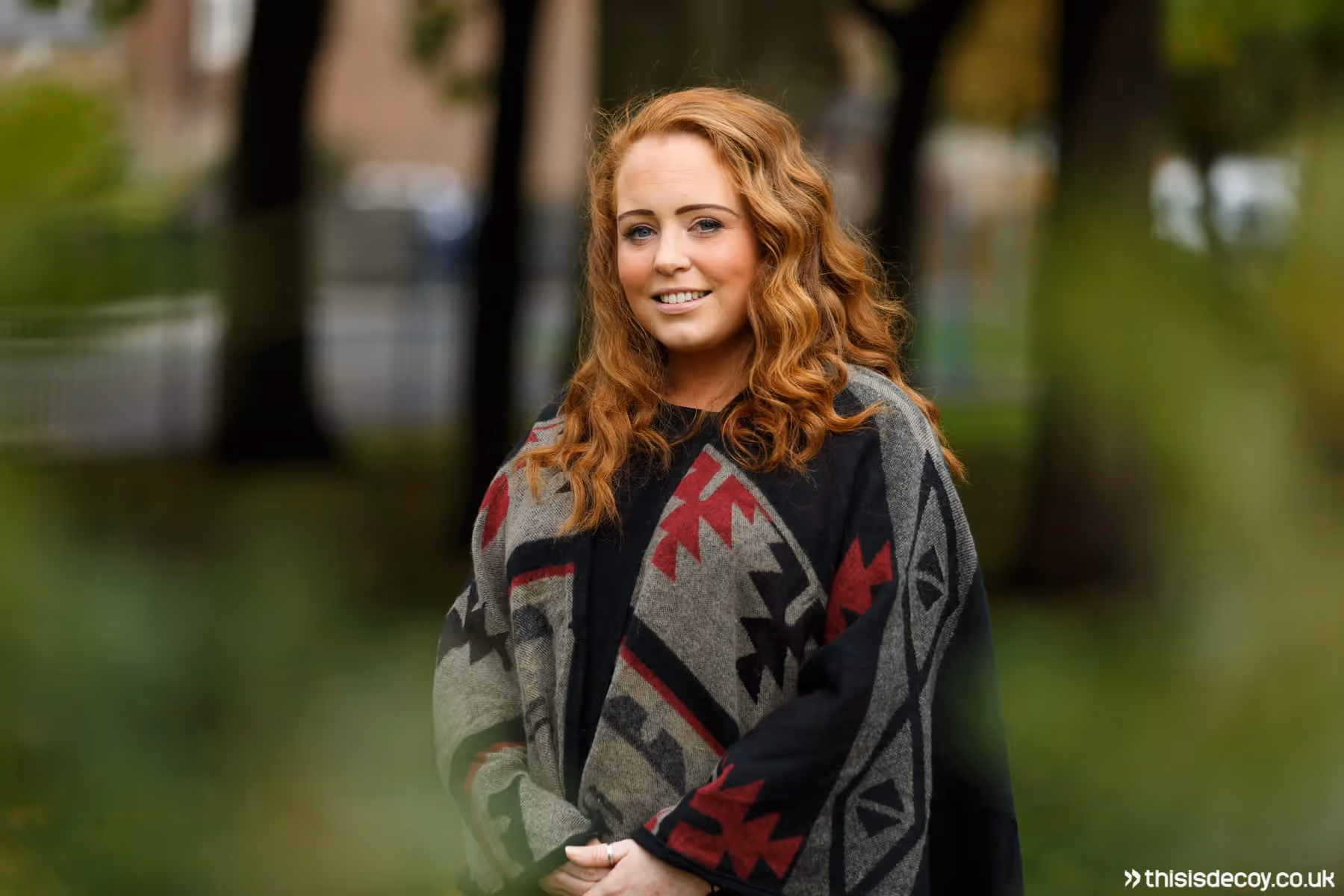 Professional portrait photograph of a young, smiling woman stood outside looking at the camera.