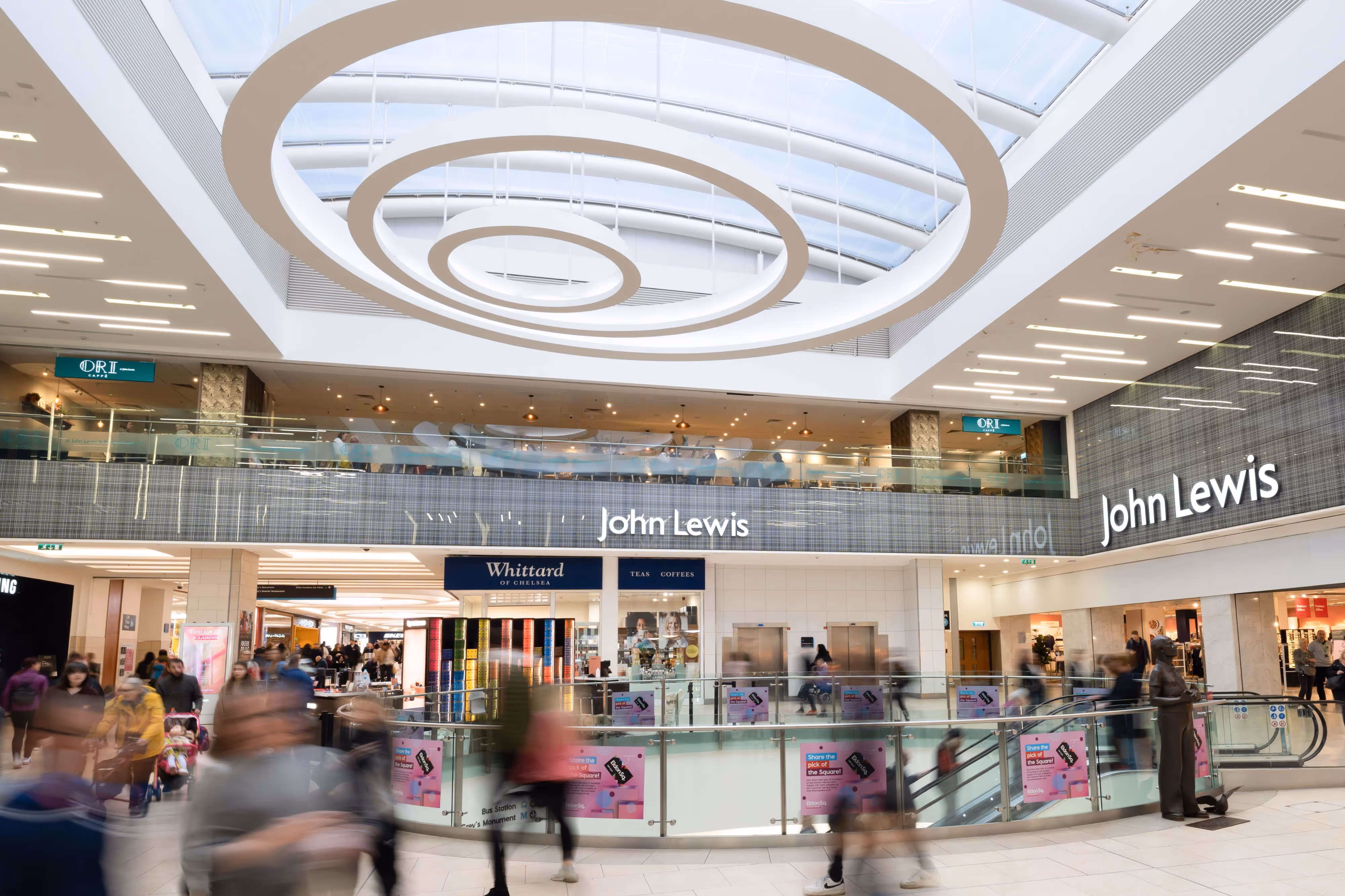 Photographer’s view of a bustling shopping centre: John Lewis shopfront, stylish circular ceiling lights, motion-blurred shoppers passing by.